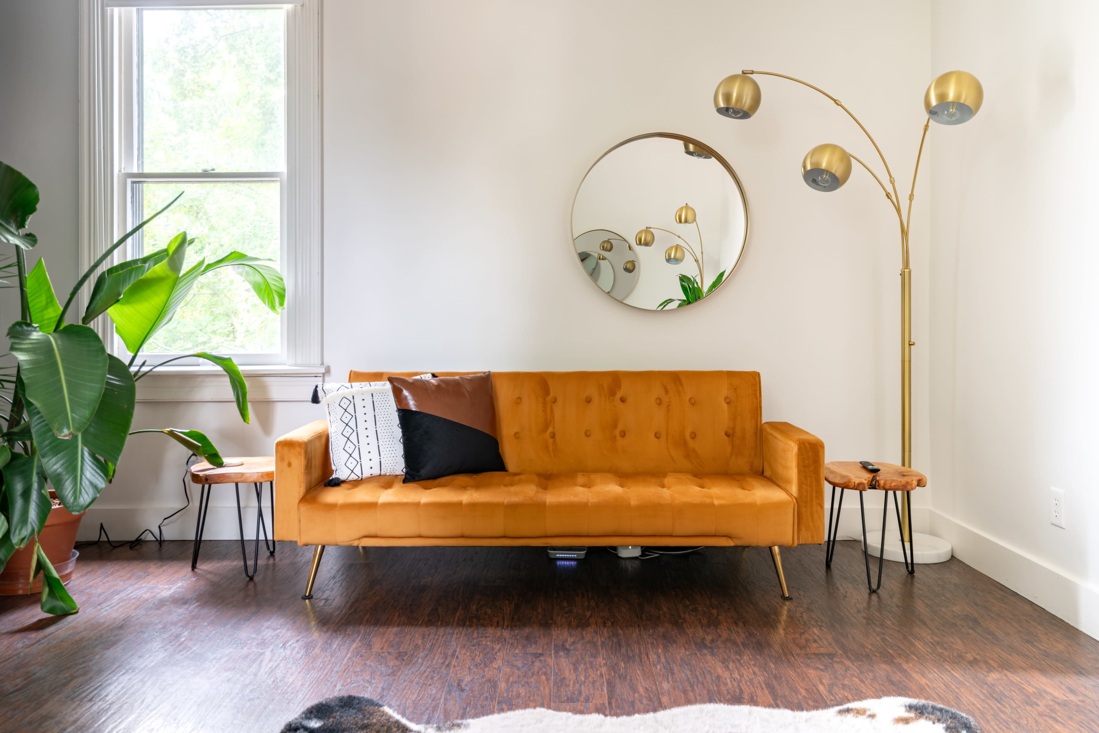 The image shows a modern living room with an orange sofa, a large round mirror, a standing lamp, and potted plants.