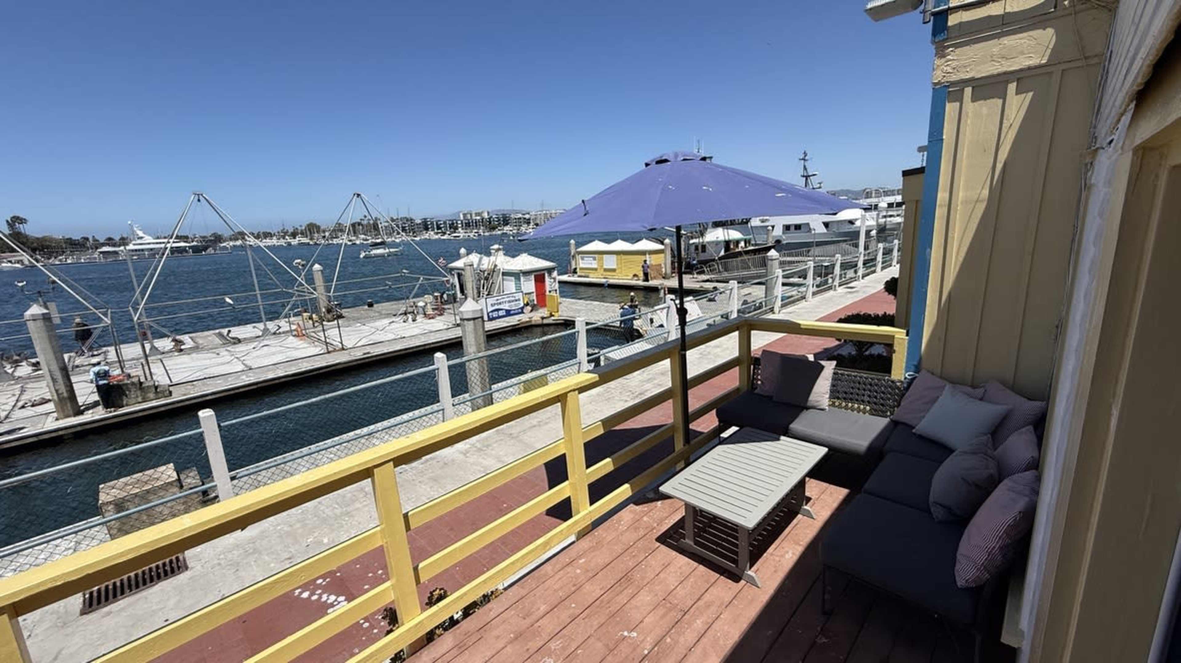 The image shows a deck with seating overlooking a marina, featuring boats docked along the water and a large umbrella in the background.