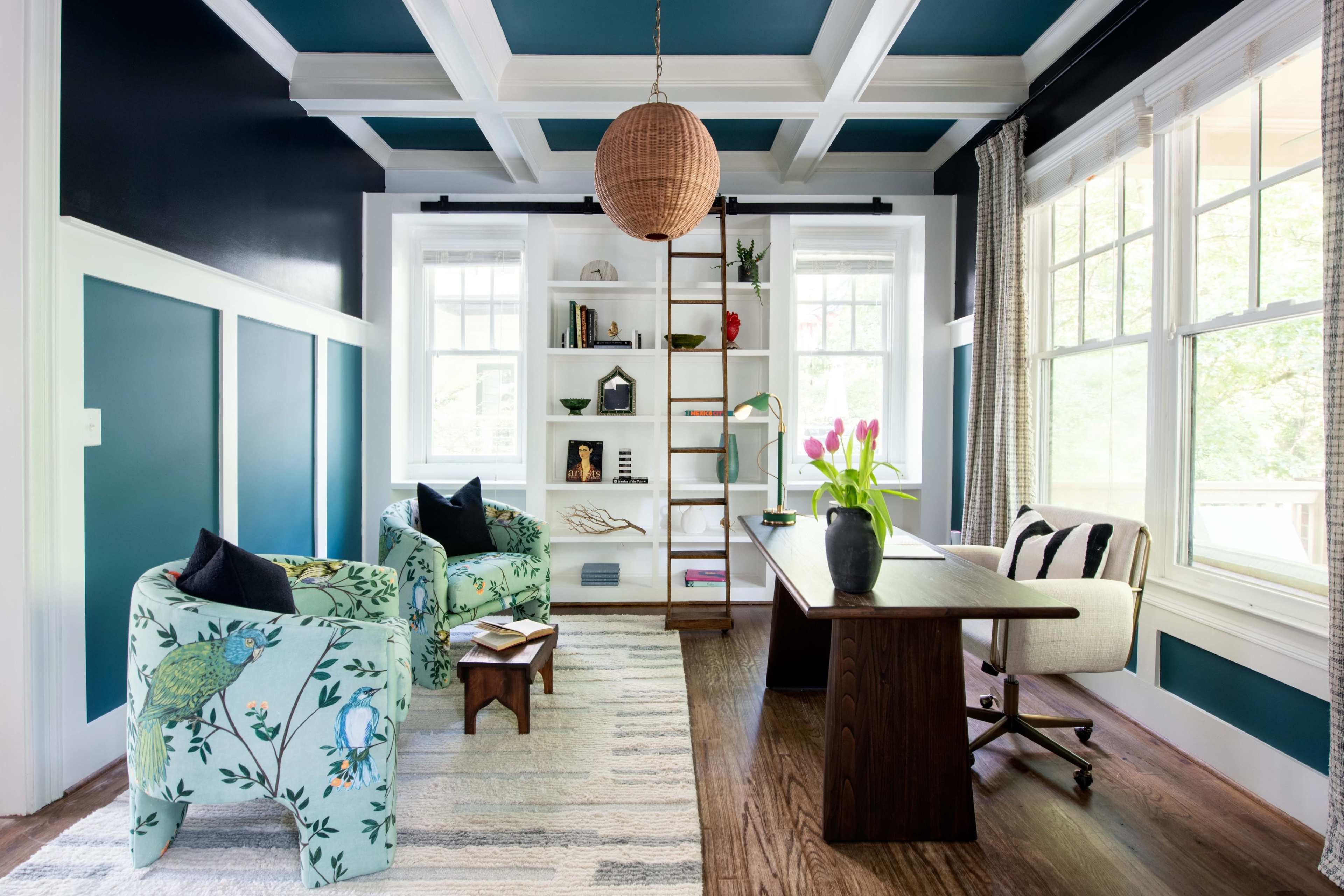 A home office features teal walls, a coffered ceiling, and a mix of floral-patterned chairs, a wooden desk, and a tall bookshelf filled with books and decorative items.