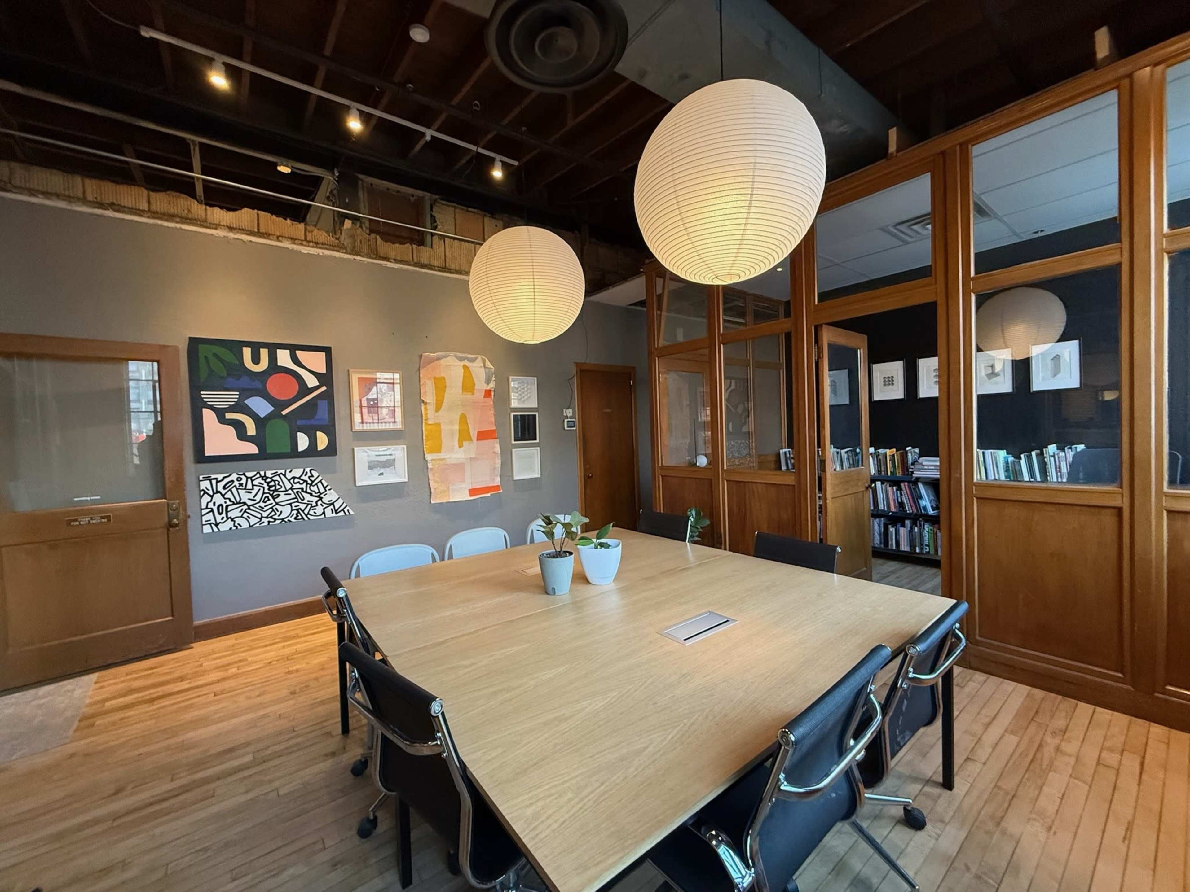 A conference room with a large wooden table, surrounded by black chairs, and illuminated by two pendant lights, features walls adorned with artwork and bookshelves in the background.