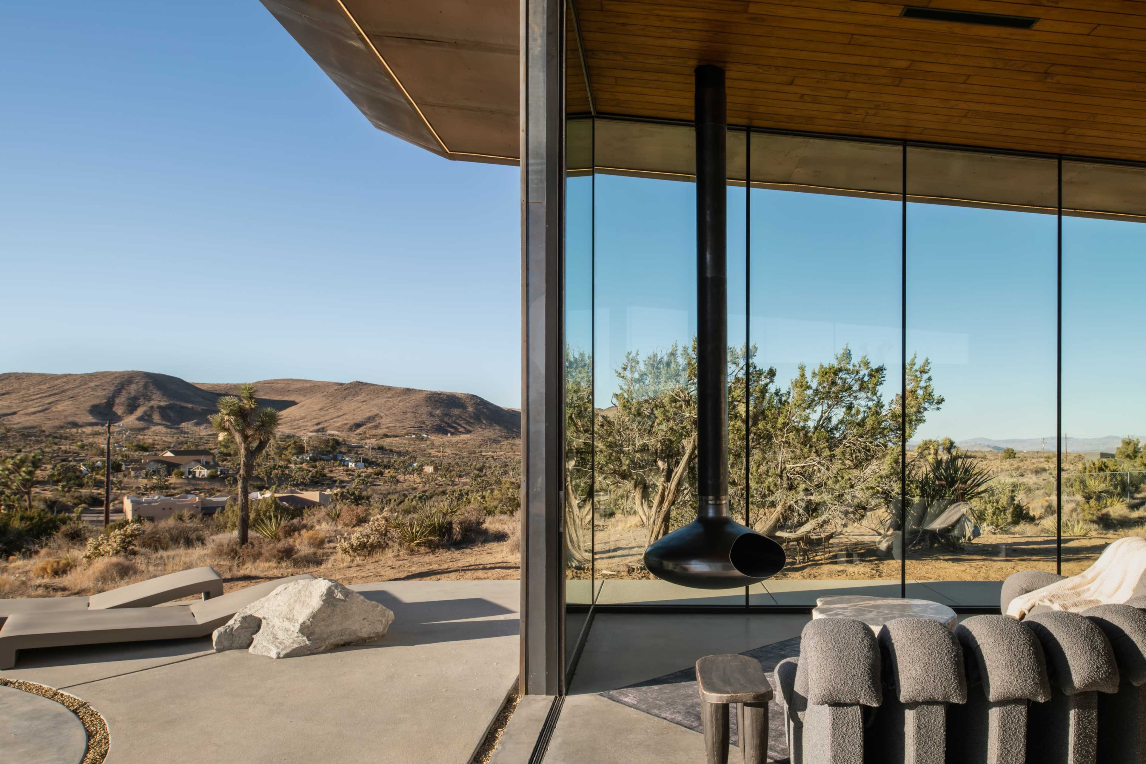 A modern glass-walled home features a central black chimney and offers a view of a desert landscape with rolling hills and scattered vegetation.