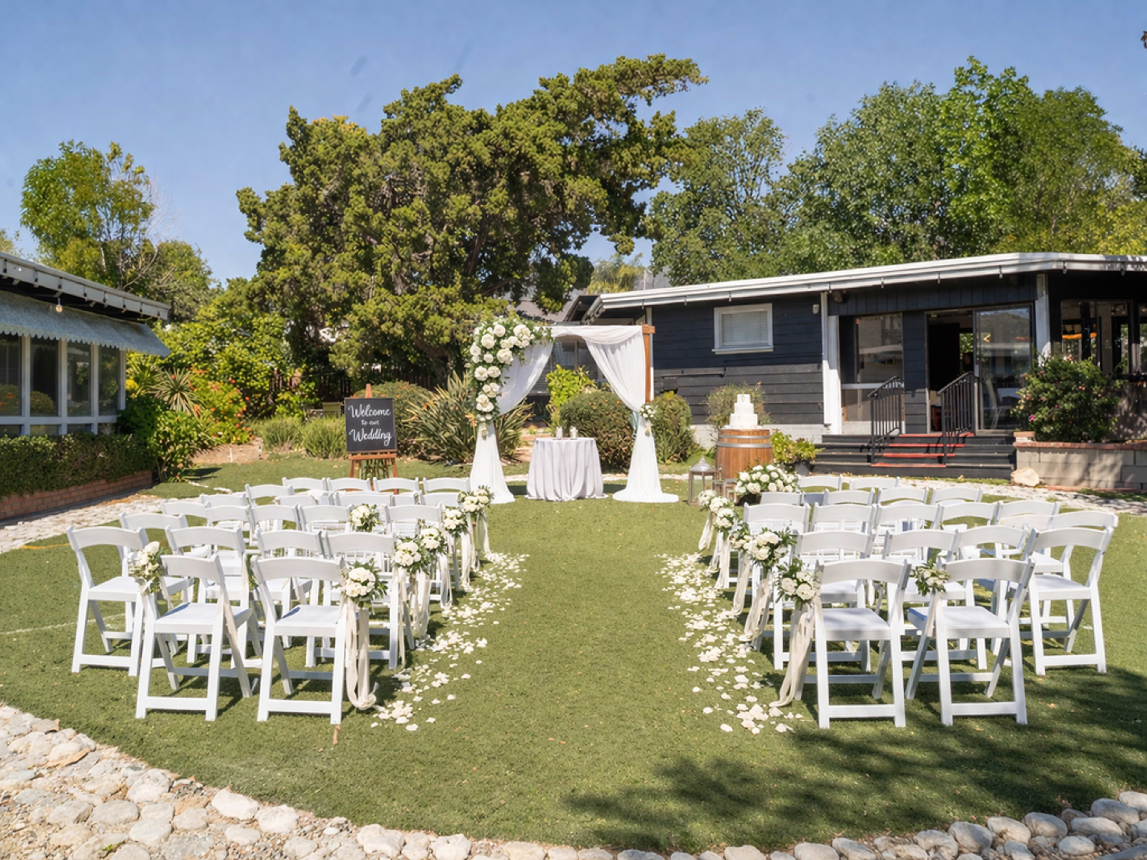 The image shows an outdoor wedding ceremony setup with white chairs arranged in rows facing an altar adorned with flowers and a wedding cake nearby.