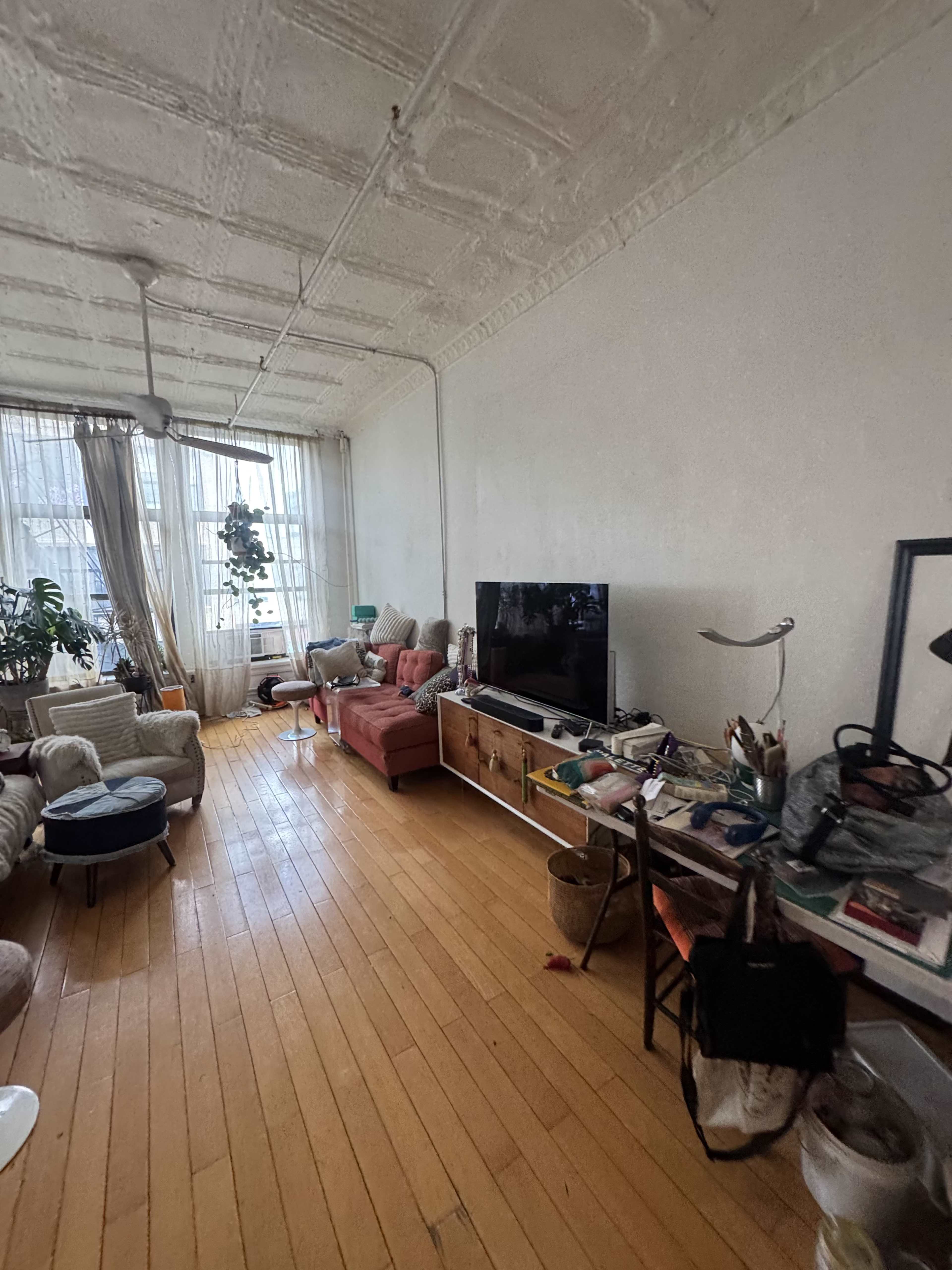 The image shows a living room with a wooden floor, a large television on a cabinet, a pink sofa, and various decorations and items scattered throughout the space.