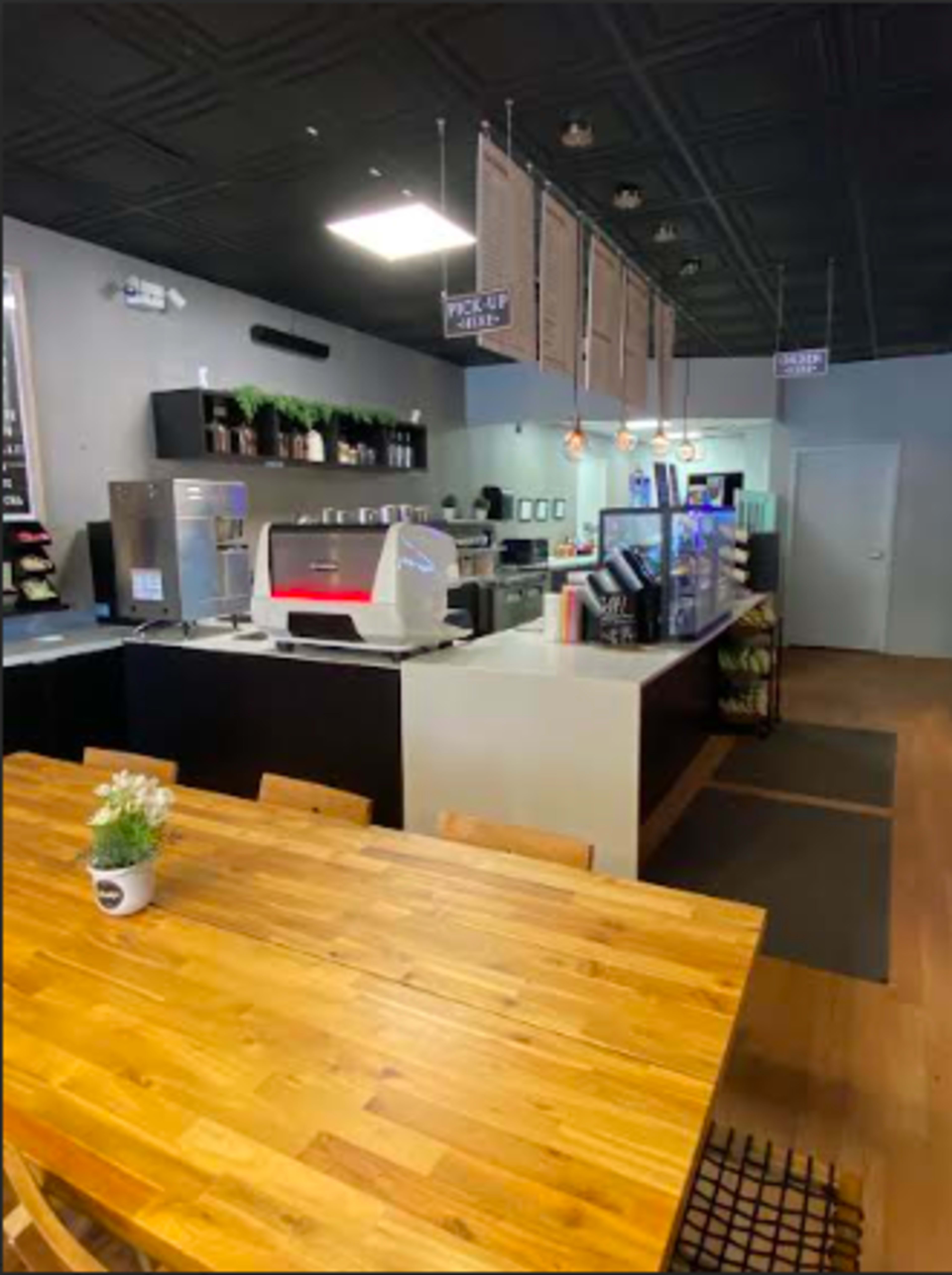 The image shows a modern café interior with a wooden table in the foreground, a beverage counter with an espresso machine, and various shelves and signs on the walls.