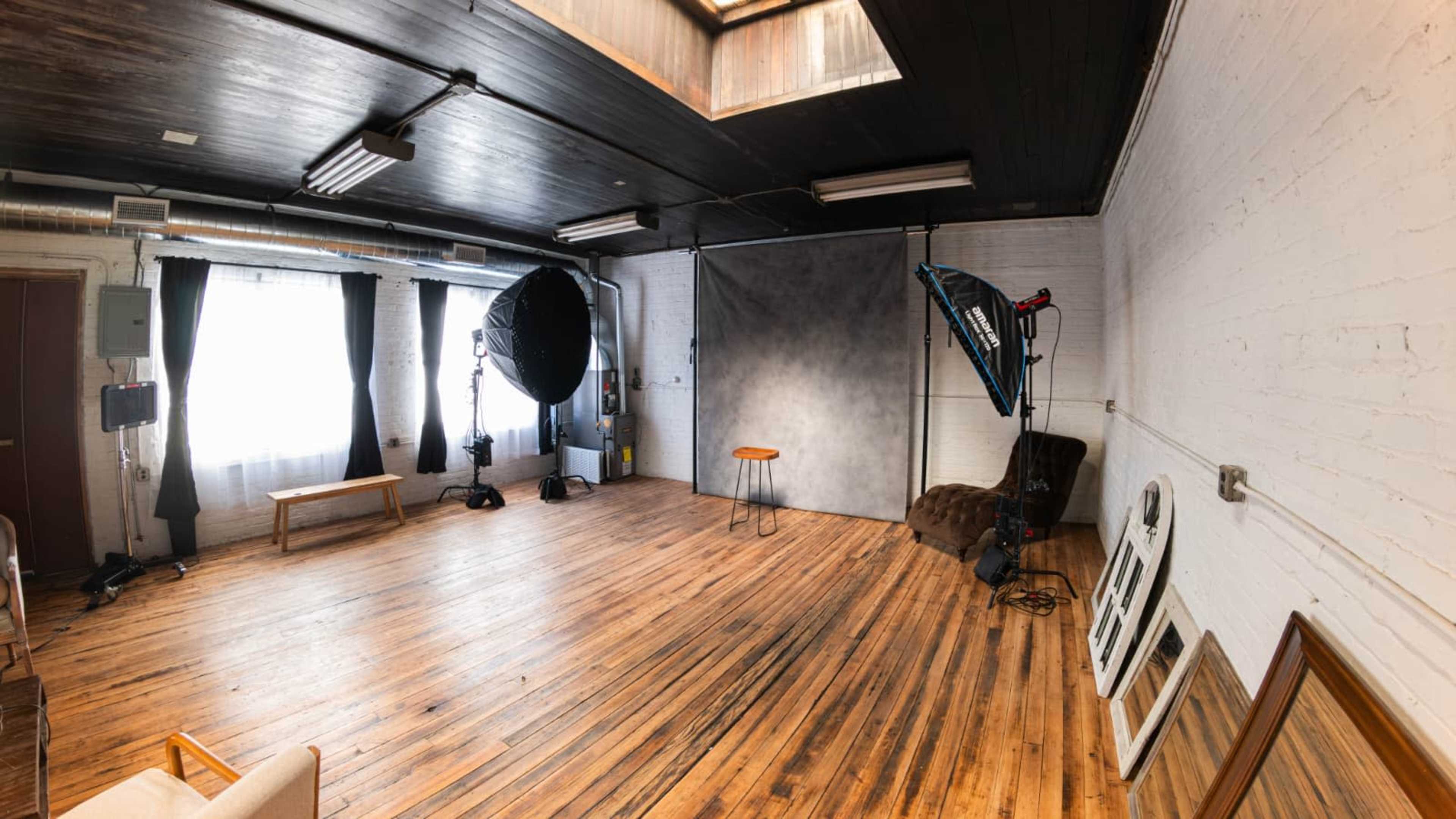 The image shows a photography studio with wooden flooring, a backdrop setup, and various lighting equipment.