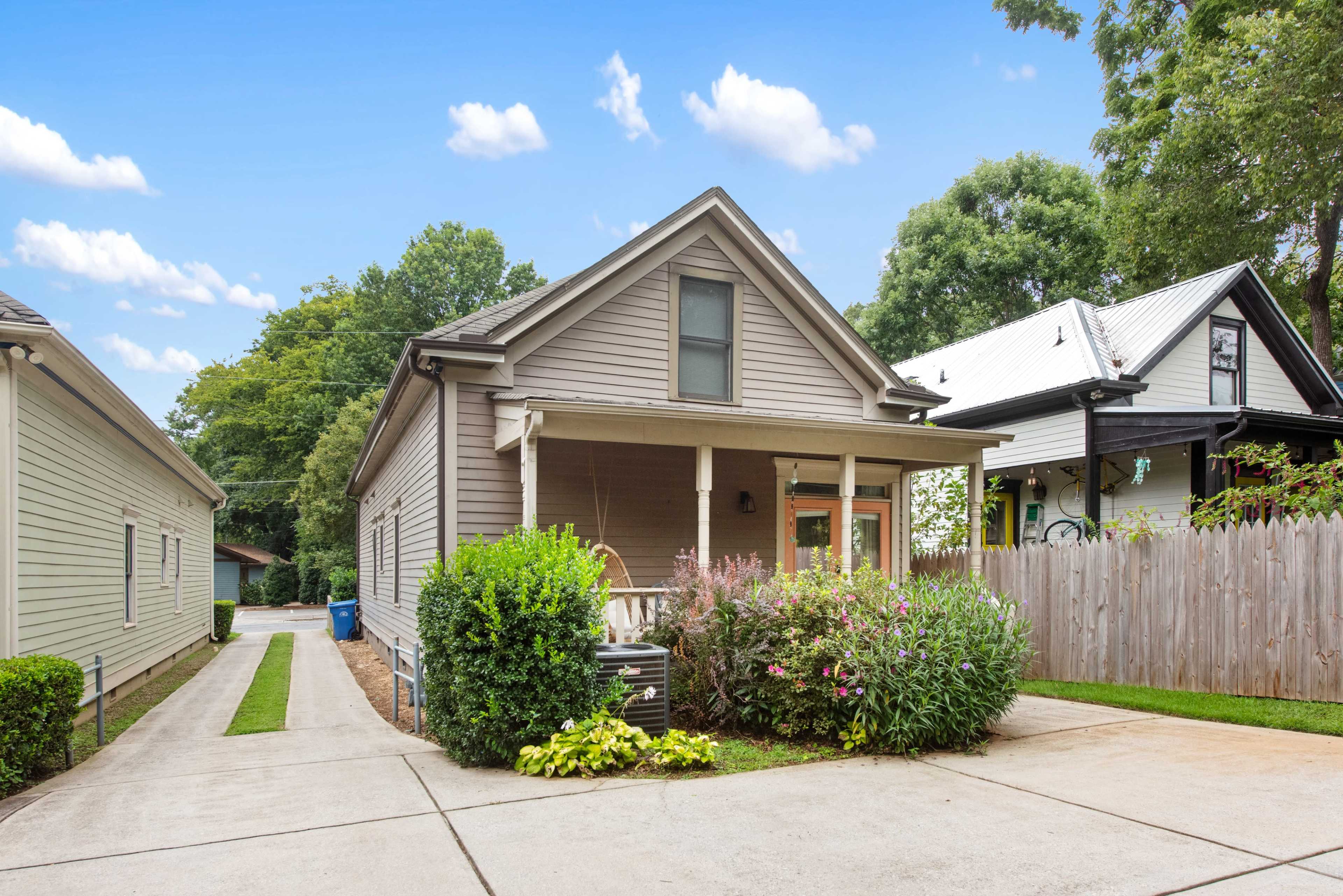 A gray house with a front porch is situated next to a narrow driveway lined with plants and flowers, surrounded by a wooden fence.