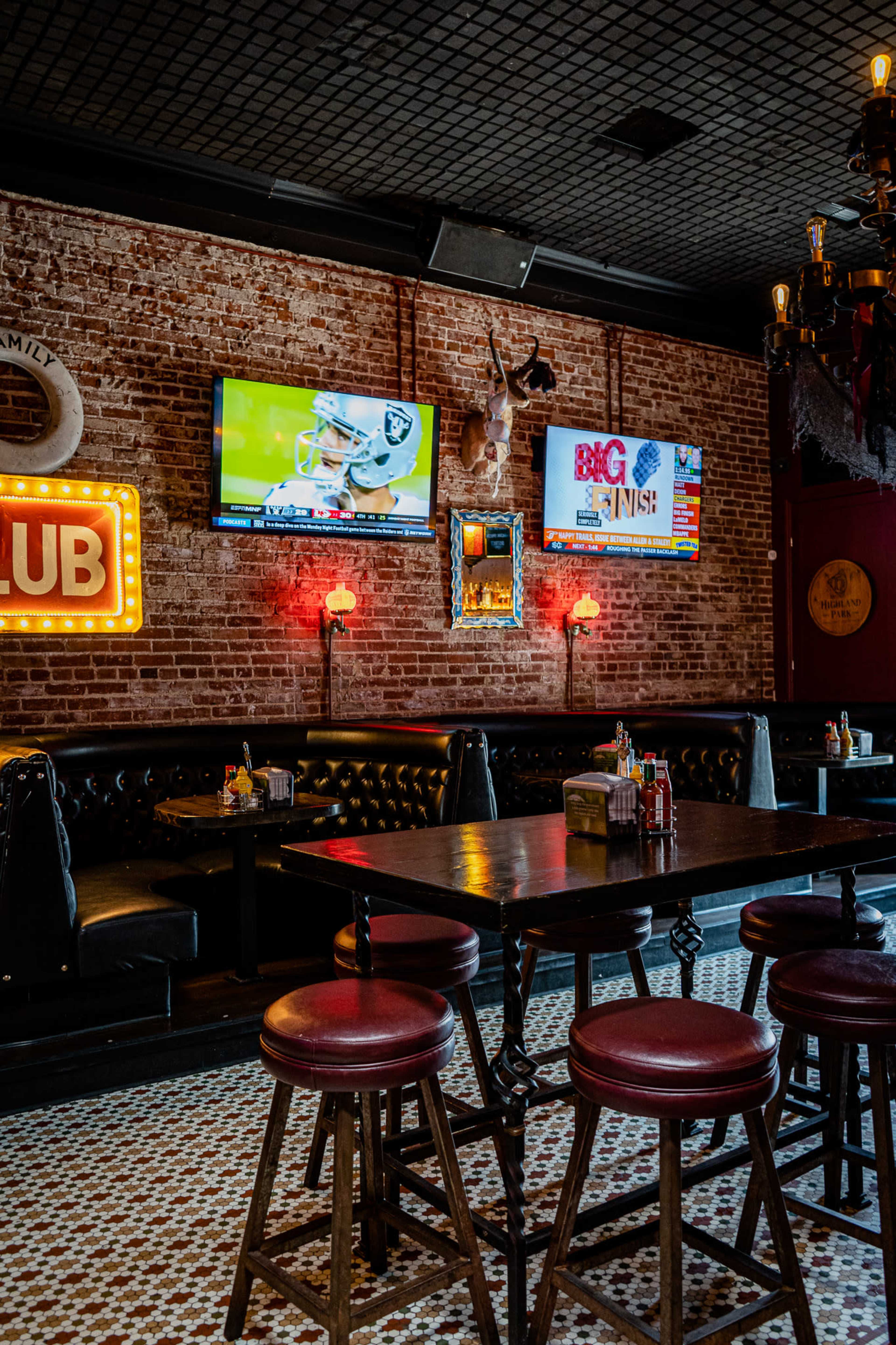 The interior of a bar features leather booths, wooden tables with red stools, and two televisions mounted on exposed brick walls.