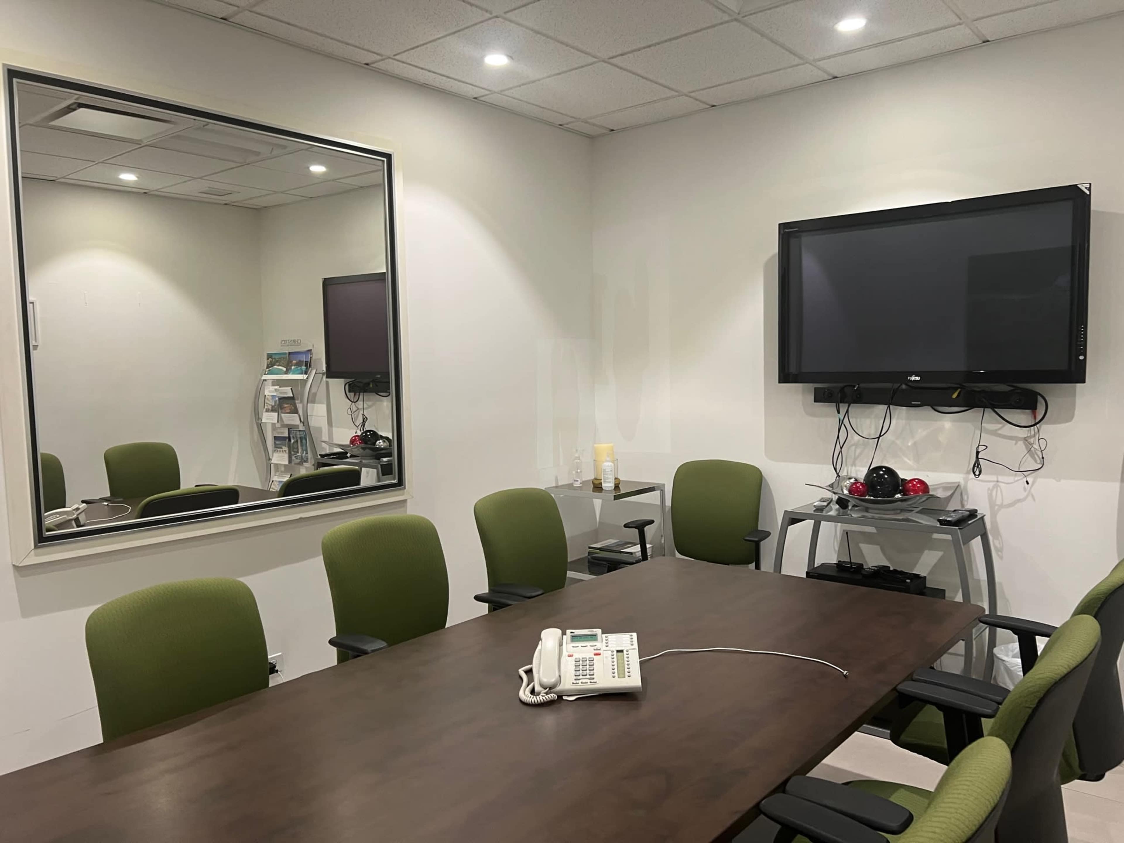 A conference room with a large table surrounded by green chairs, a wall-mounted television, a phone on the table, and a mirror reflecting the room.