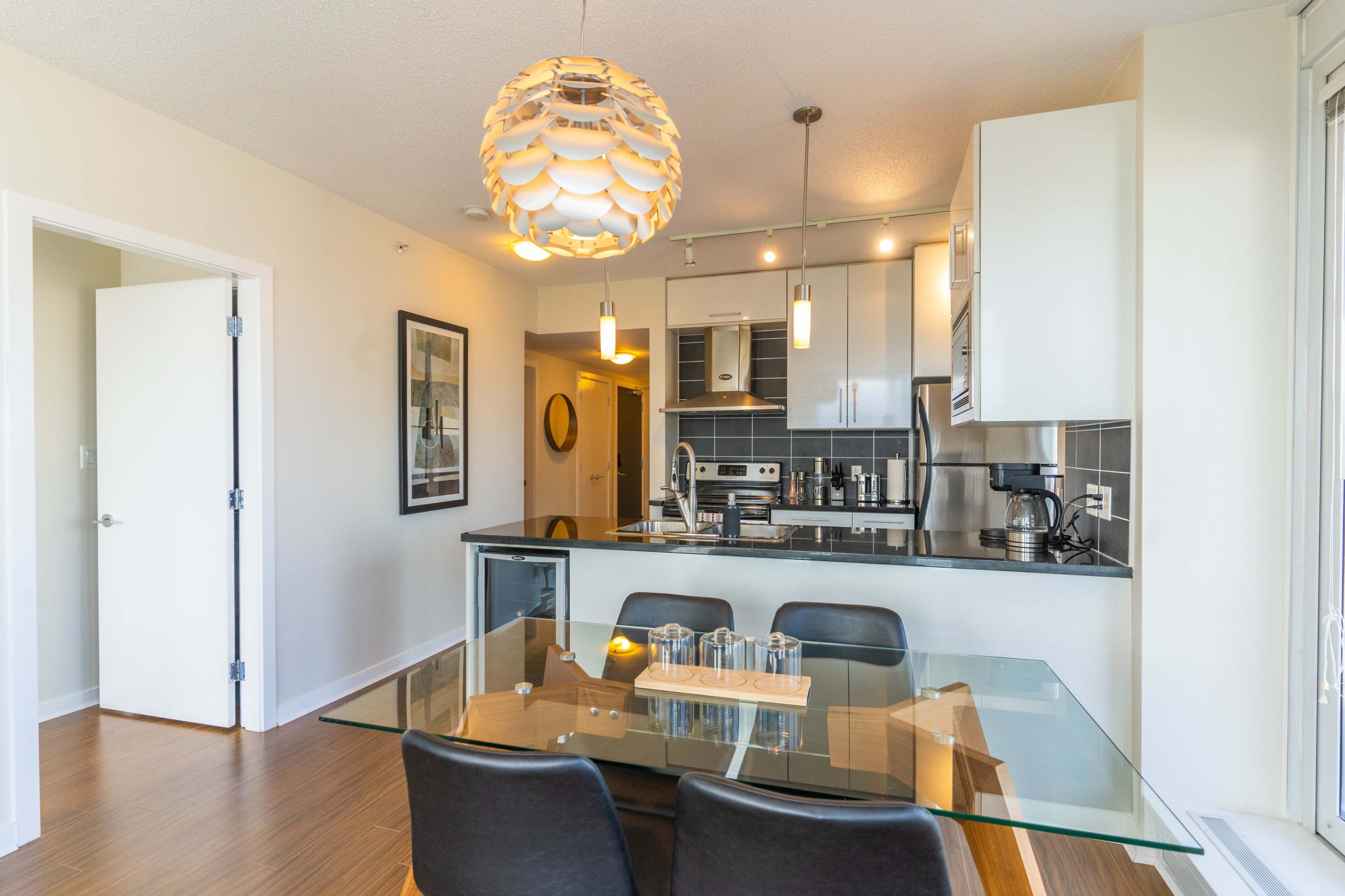 A modern kitchen with a glass dining table and four black chairs, featuring white cabinets and stainless steel appliances.