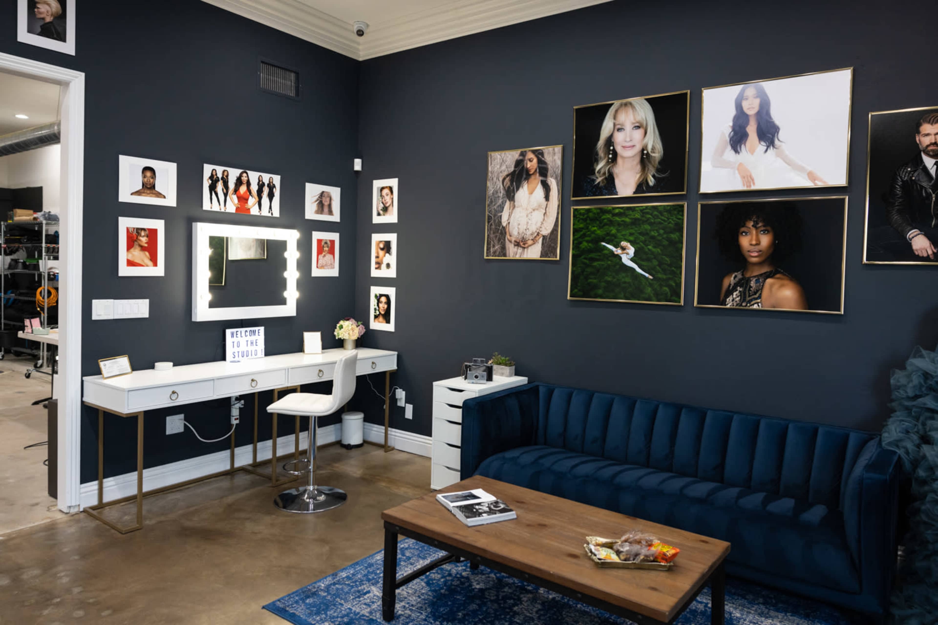 A modern beauty studio featuring a blue velvet sofa, a white vanity with a lighted mirror, and several framed portraits on the dark wall.