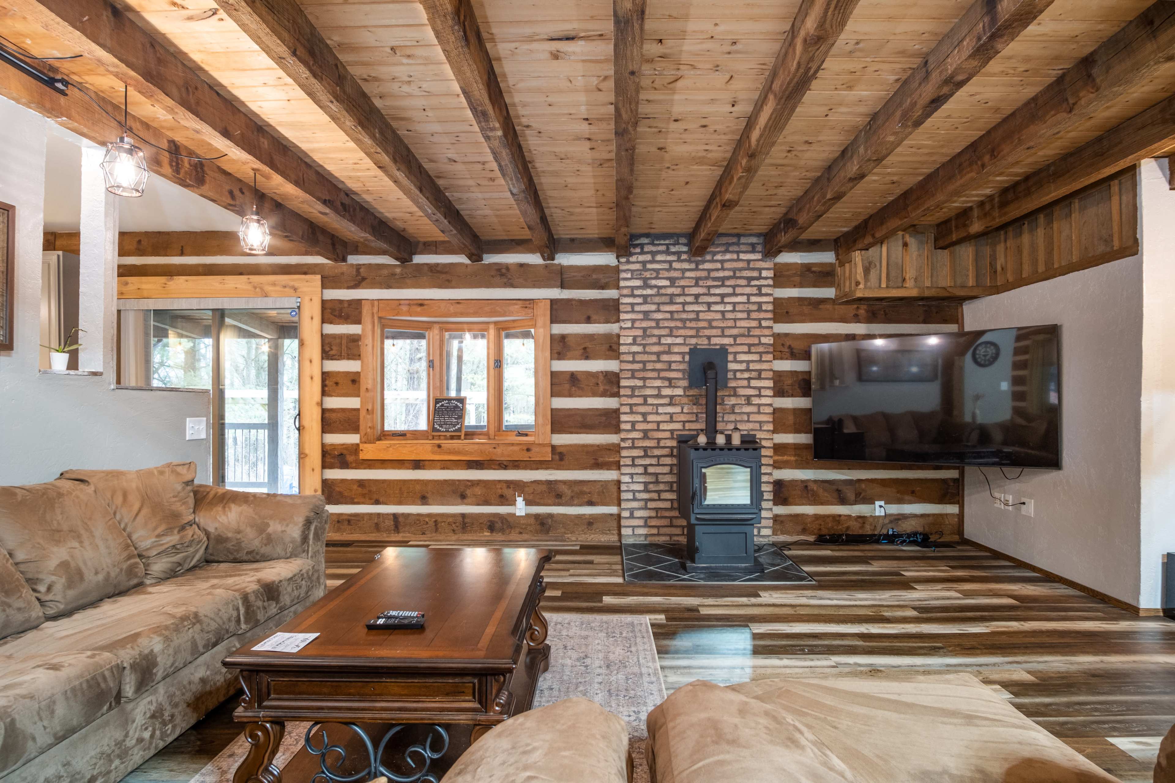 The interior features a wooden-beamed ceiling, a brick chimney, a dark sofa, and a television mounted on a rustic wall.
