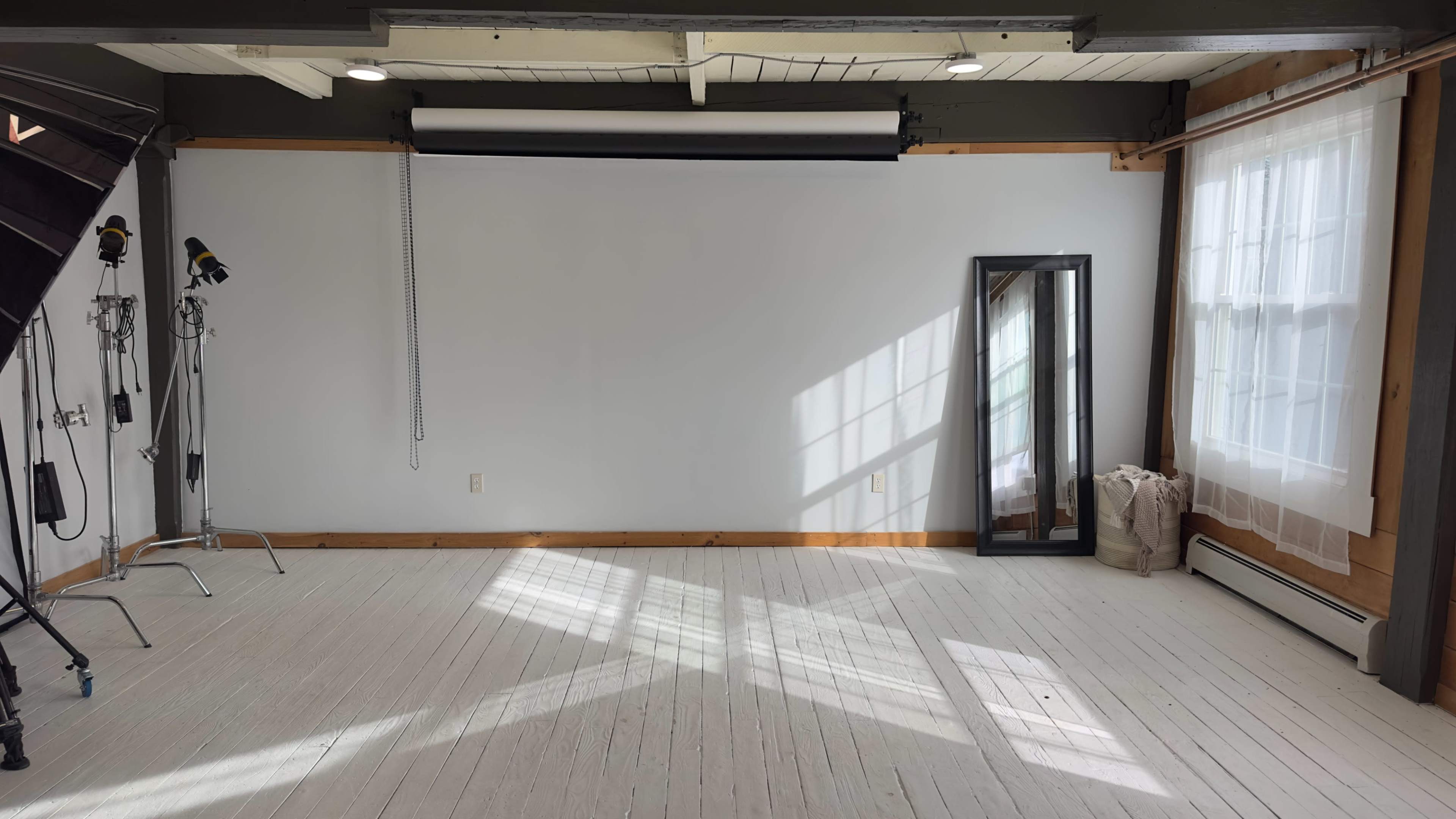 The image shows a spacious room with a gray wall, wooden accents, a large mirror, and sunlight casting shadows across the white wooden floor.