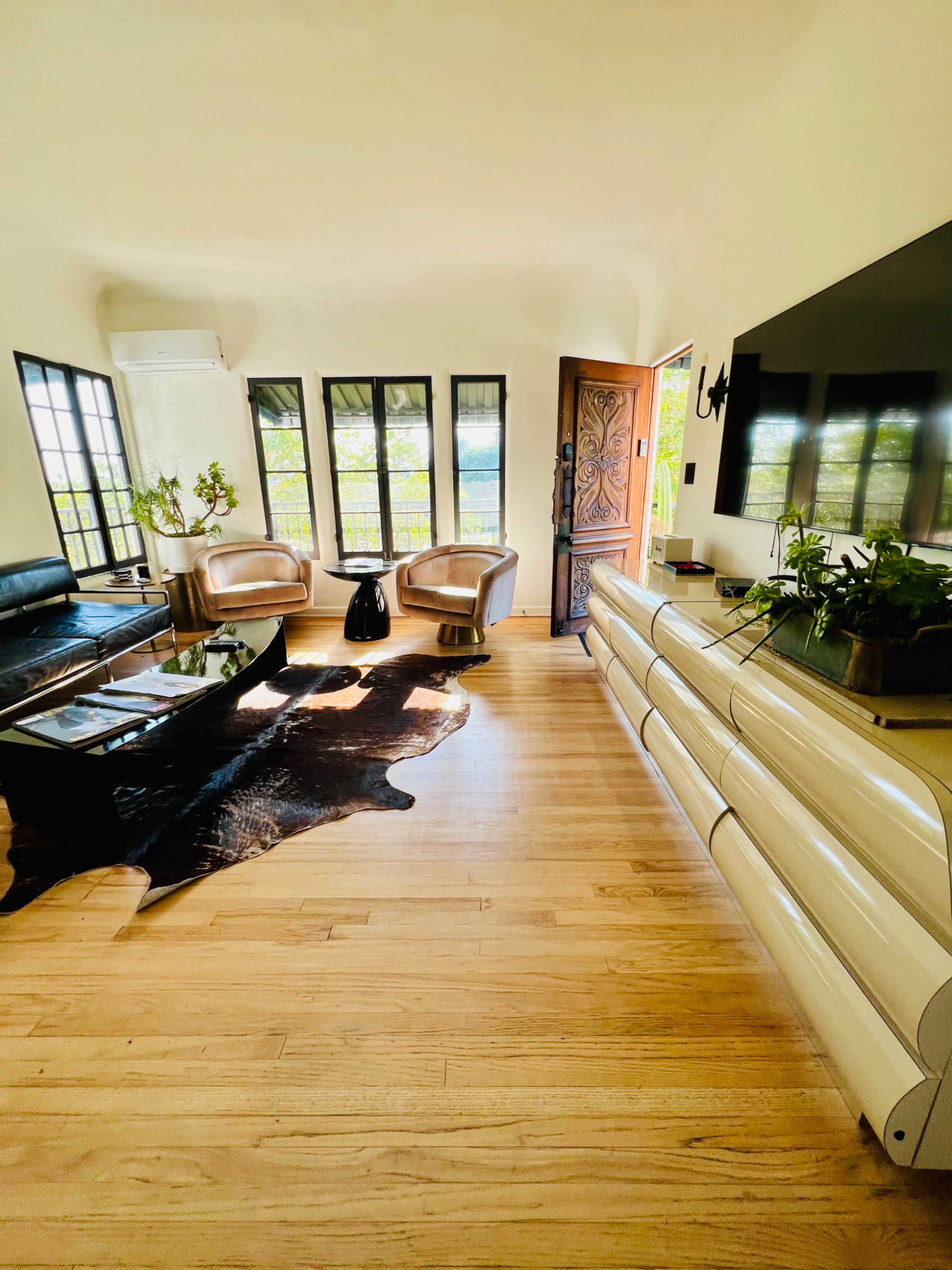 The image shows a bright living room with wooden floors, a black leather sofa, two light-colored armchairs, a glass coffee table, and large windows allowing natural light.