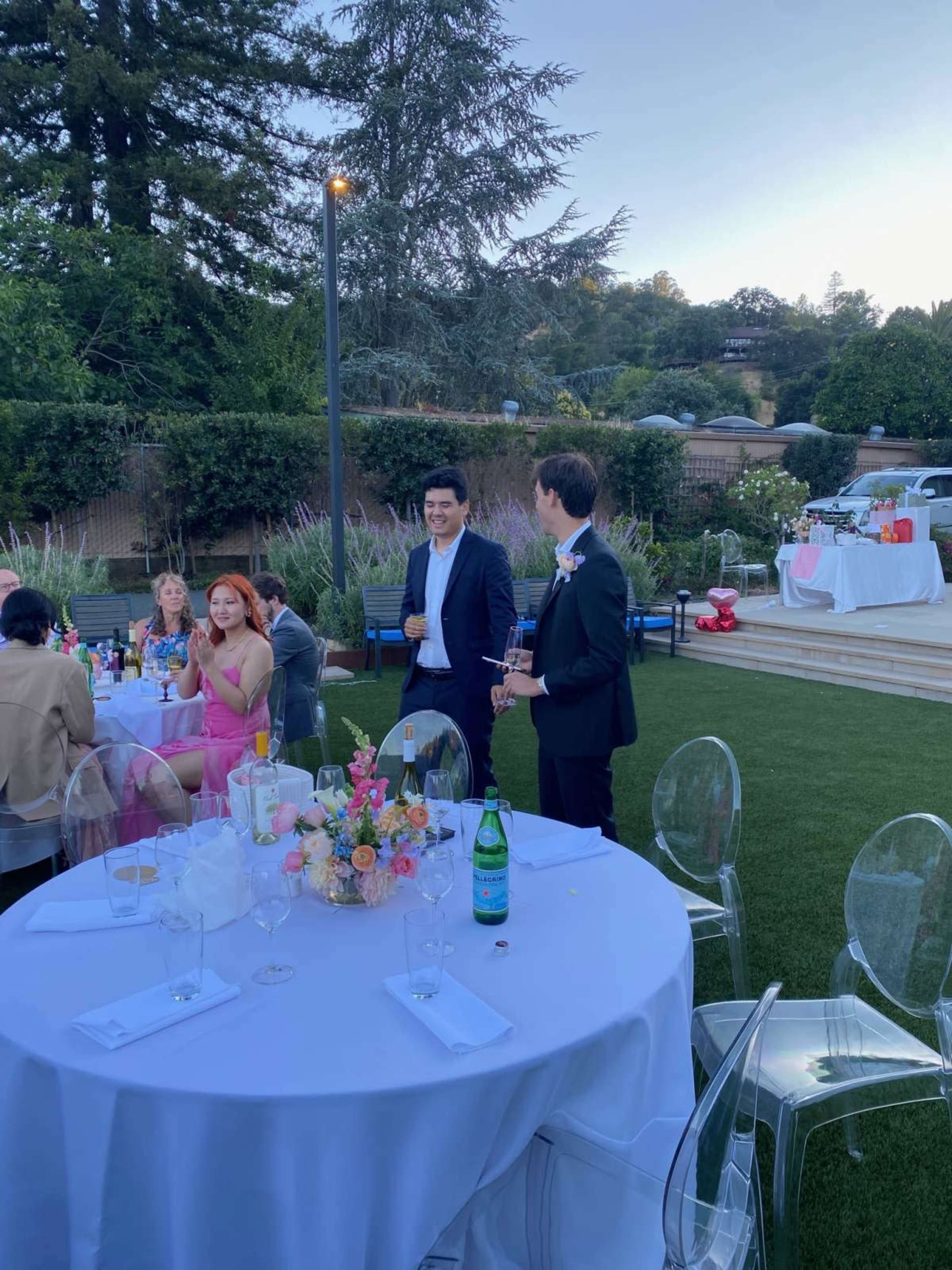 A group of people are gathered outdoors for a celebration, with tables set and refreshments on display in the background.