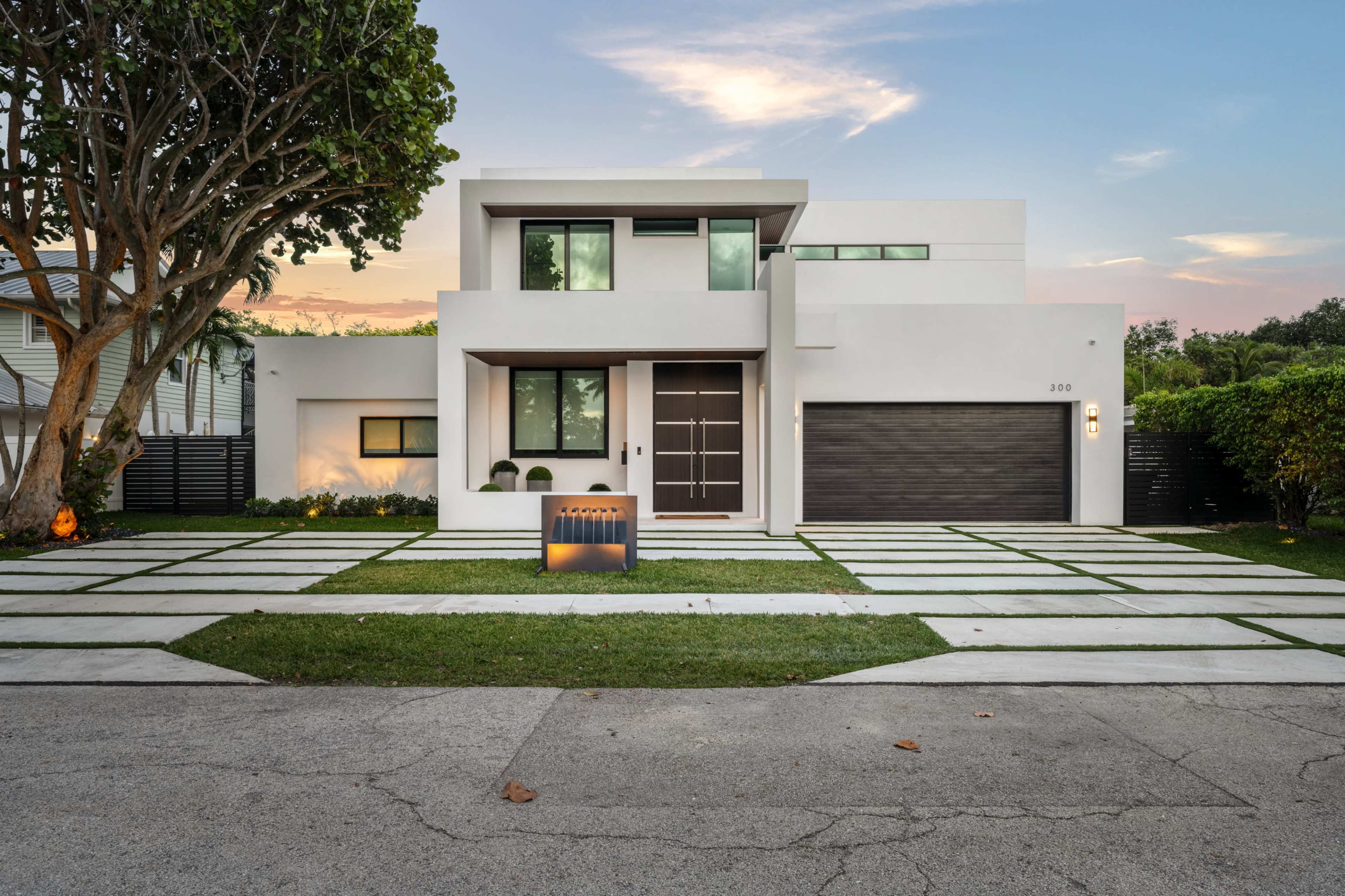 A modern two-story house with a clean white facade, large windows, and a garage, set on a neatly landscaped yard with geometric pathways.
