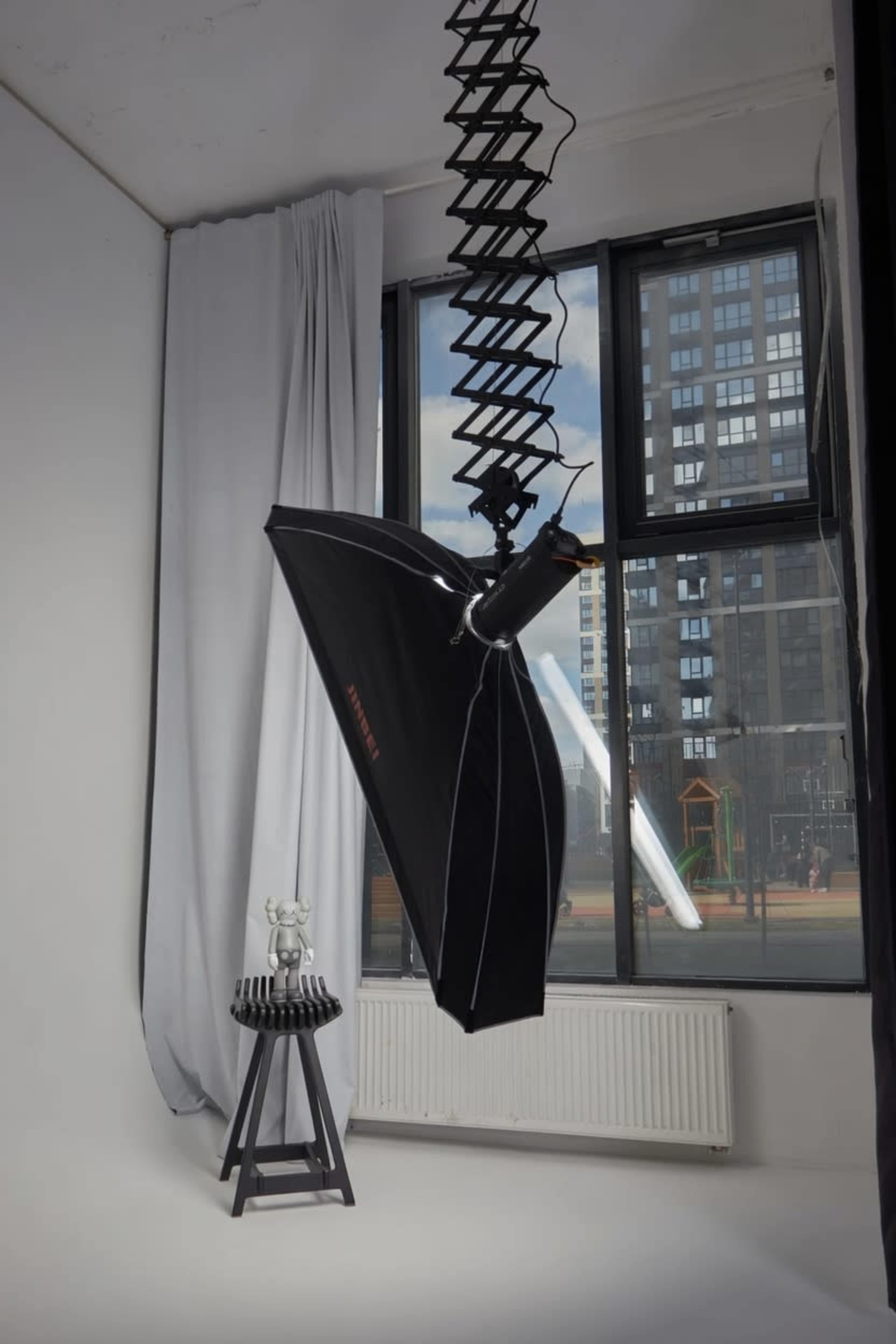 A large softbox light hangs from the ceiling above a small black table with unique decorative items near a window overlooking an urban landscape.