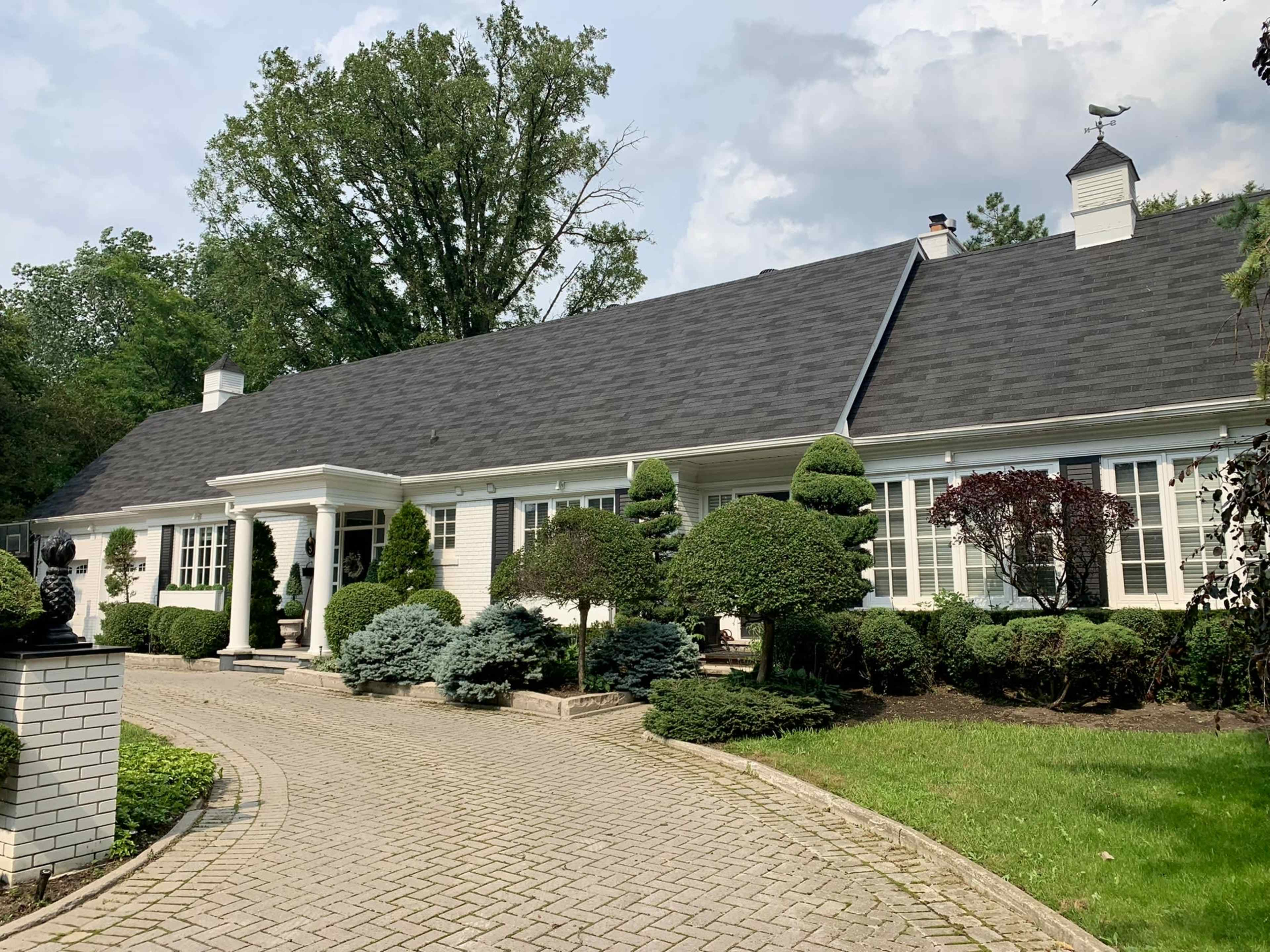 The image shows a large, single-story house surrounded by neatly trimmed bushes and trees, with a brick pathway leading to the entrance.