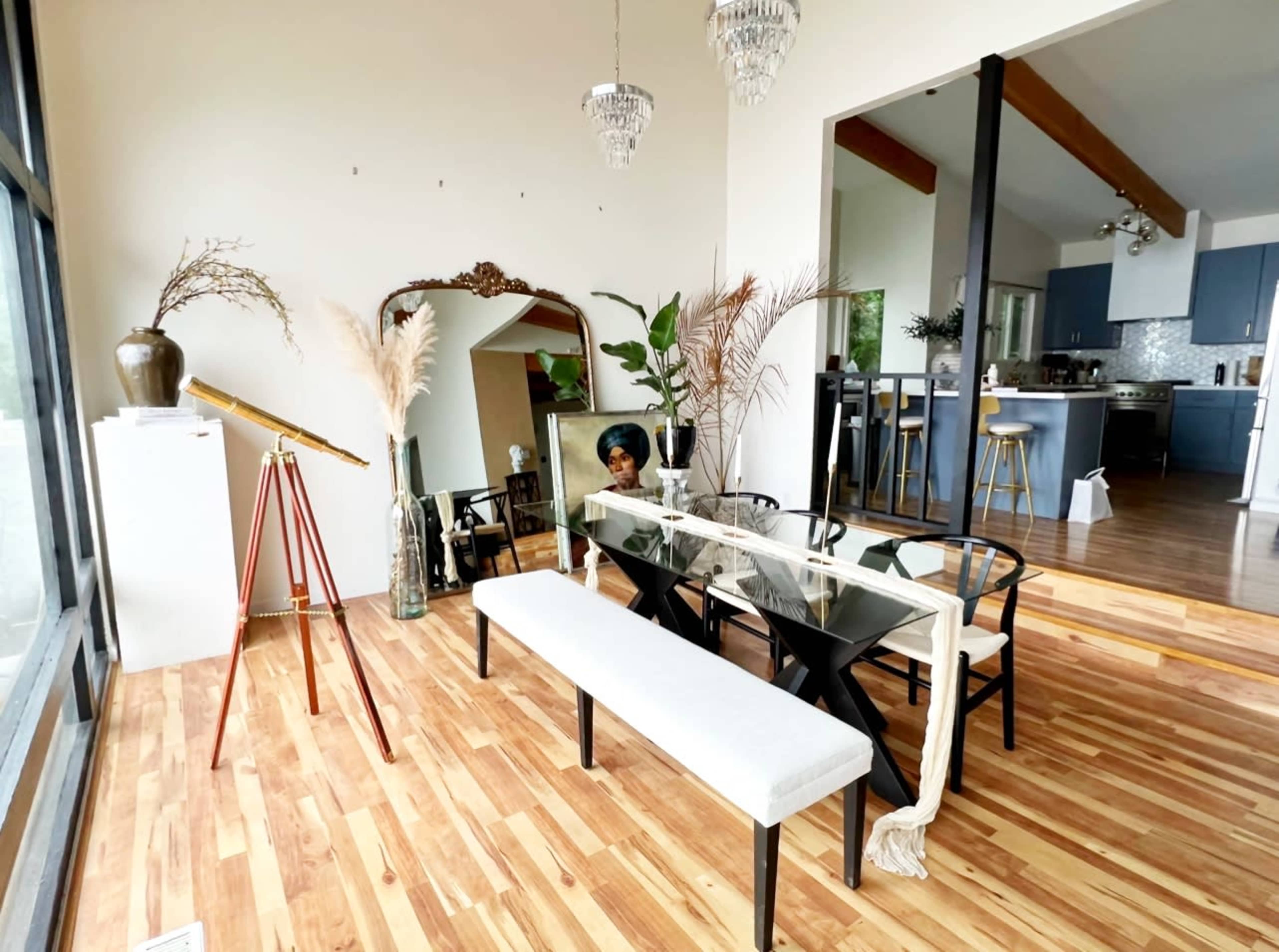 A bright dining area features a long table with black chairs, a bench, a telescope, and plants, with a kitchen visible in the background.