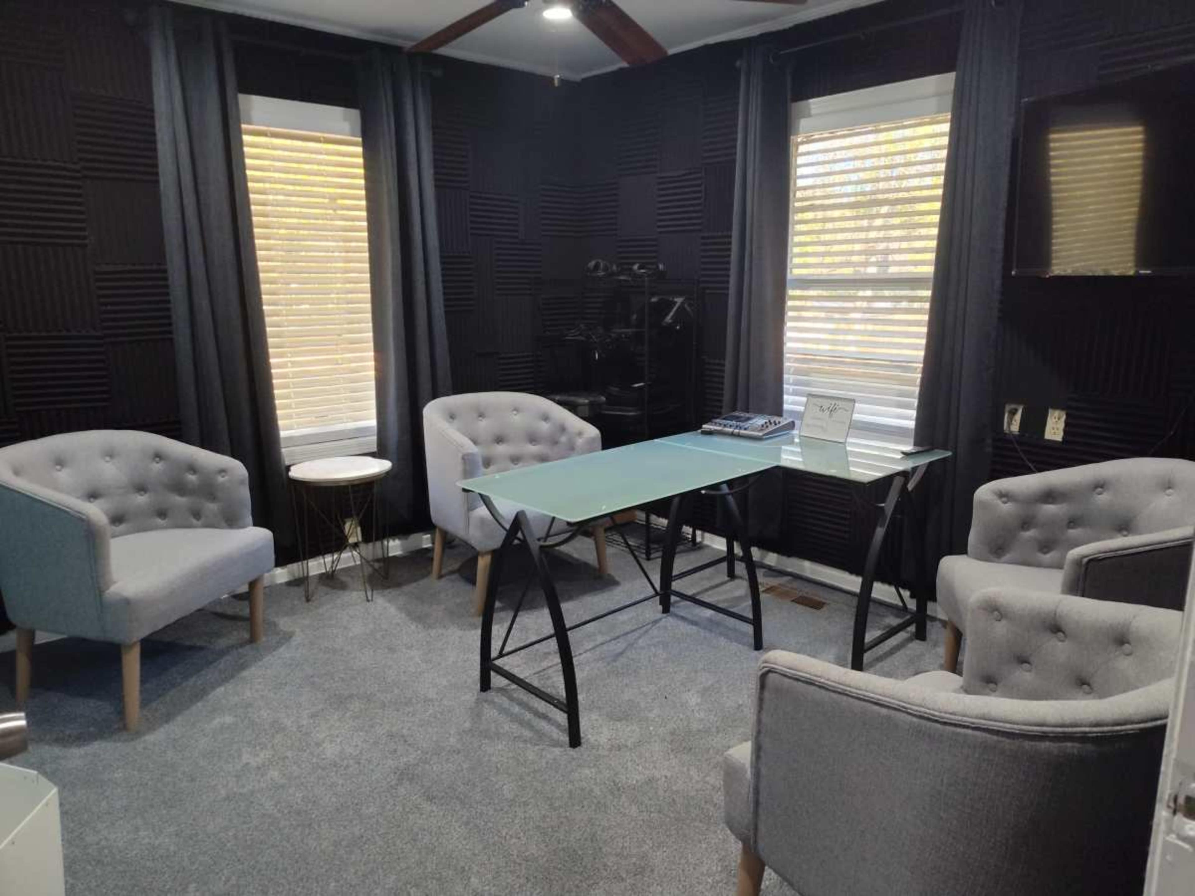 The image shows a small room with two windows, grey upholstered chairs arranged around a glass-top desk, and soundproofing panels on the walls.