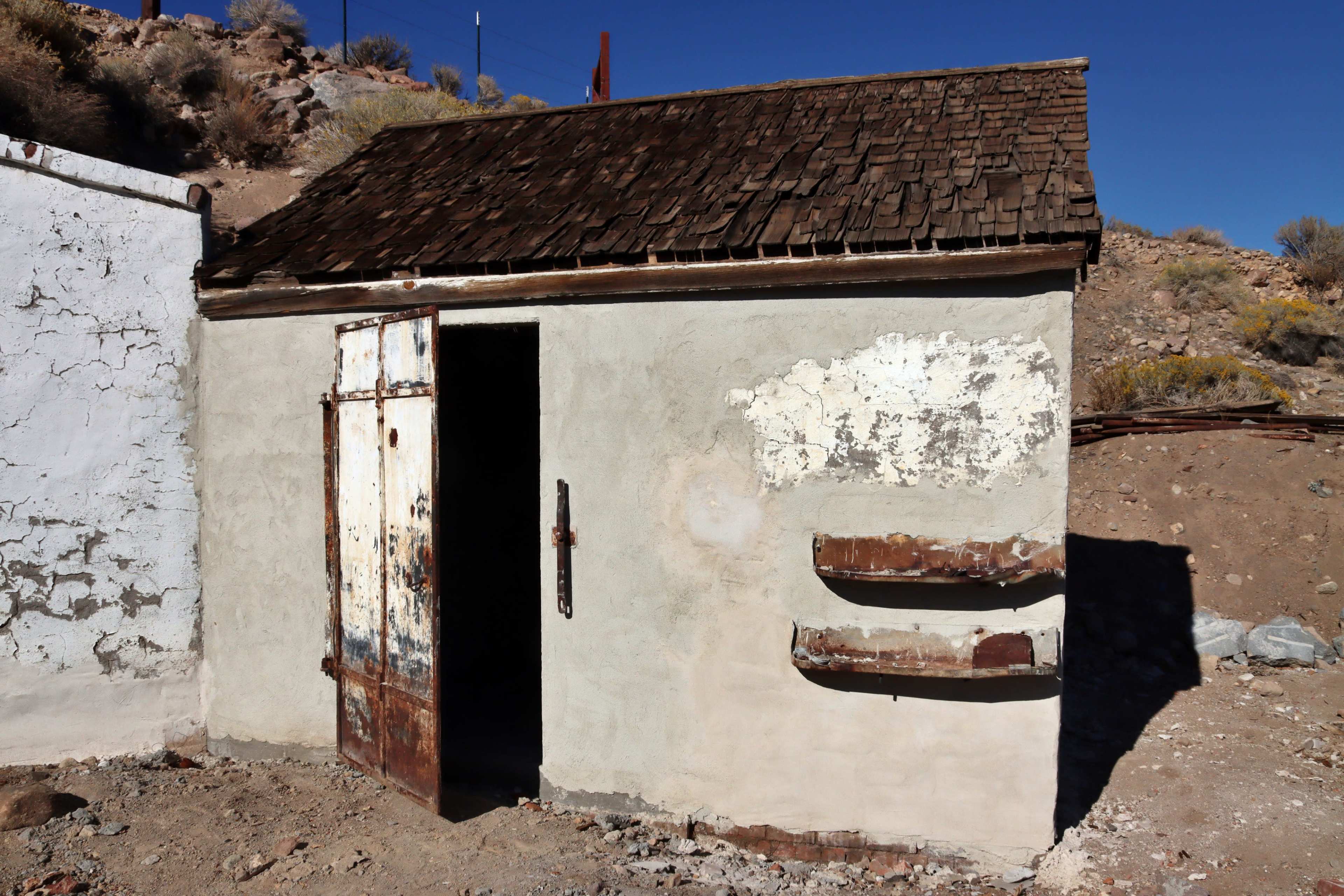 A small, weathered building with a wooden roof and a rusty metal door in a barren landscape.