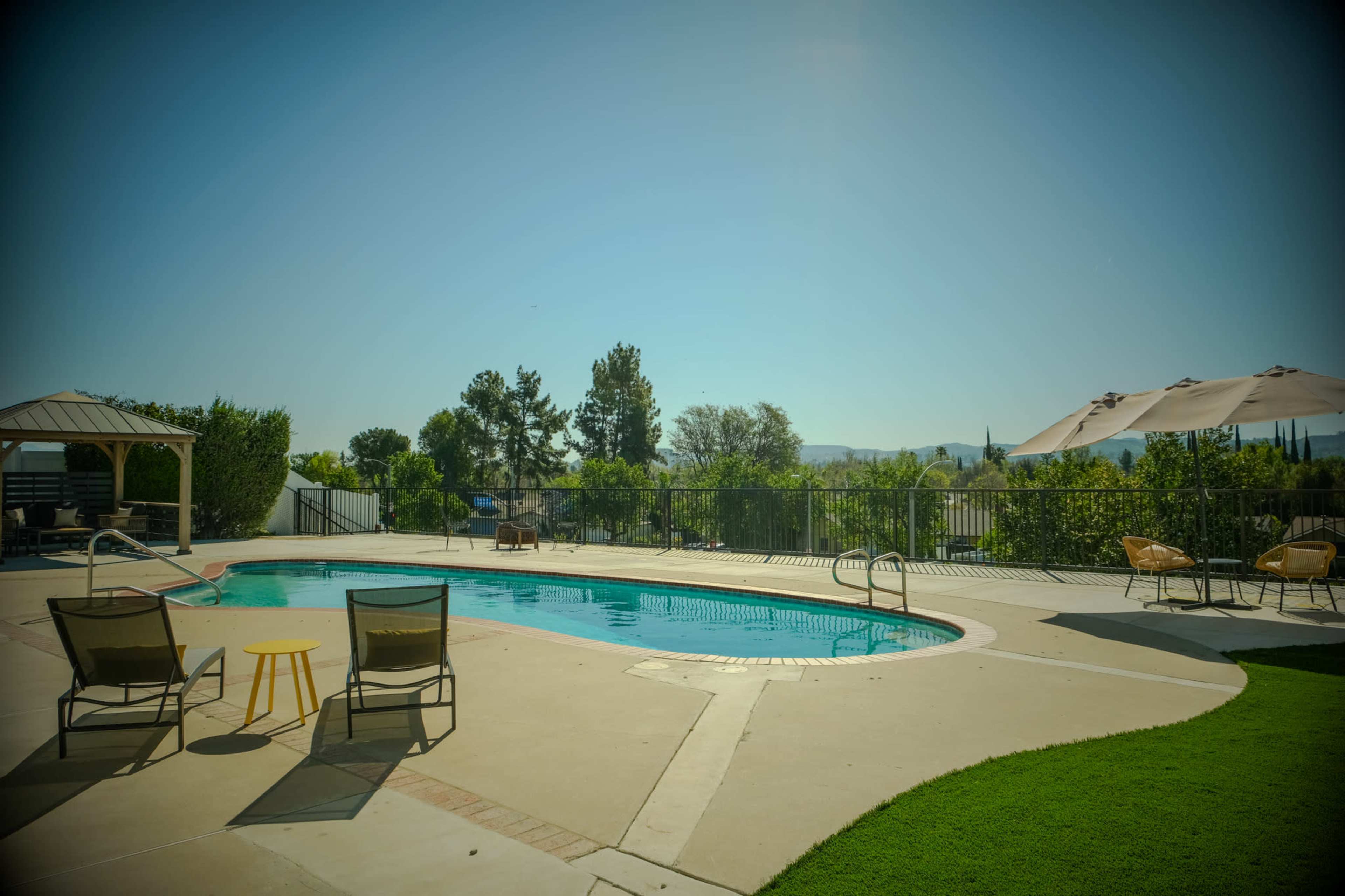 The image shows a swimming pool area surrounded by lounge chairs, a gazebo, and green landscaping under a clear blue sky.