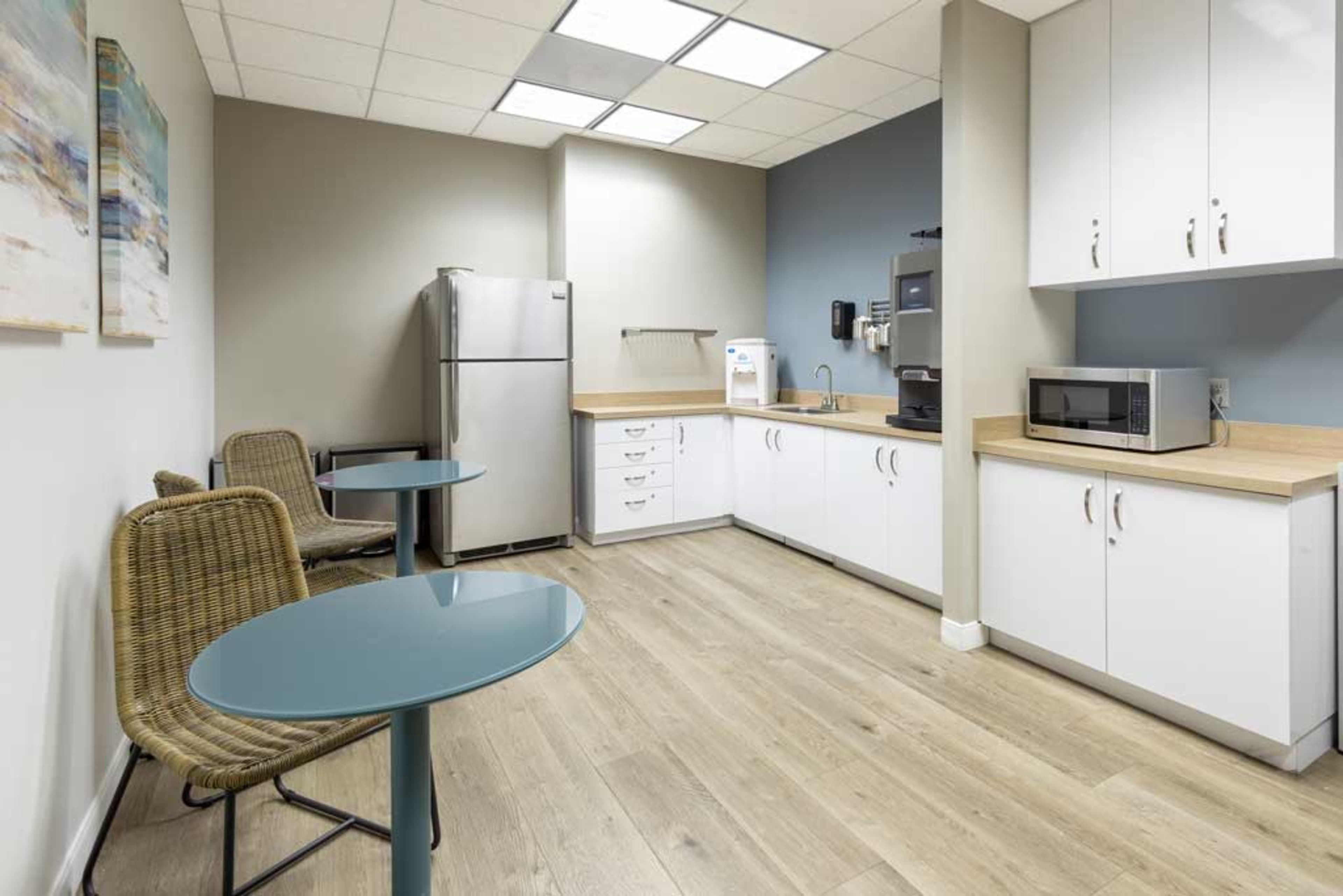 A modern break room with a refrigerator, microwave, coffee machine, and seating areas featuring two small tables and chairs.