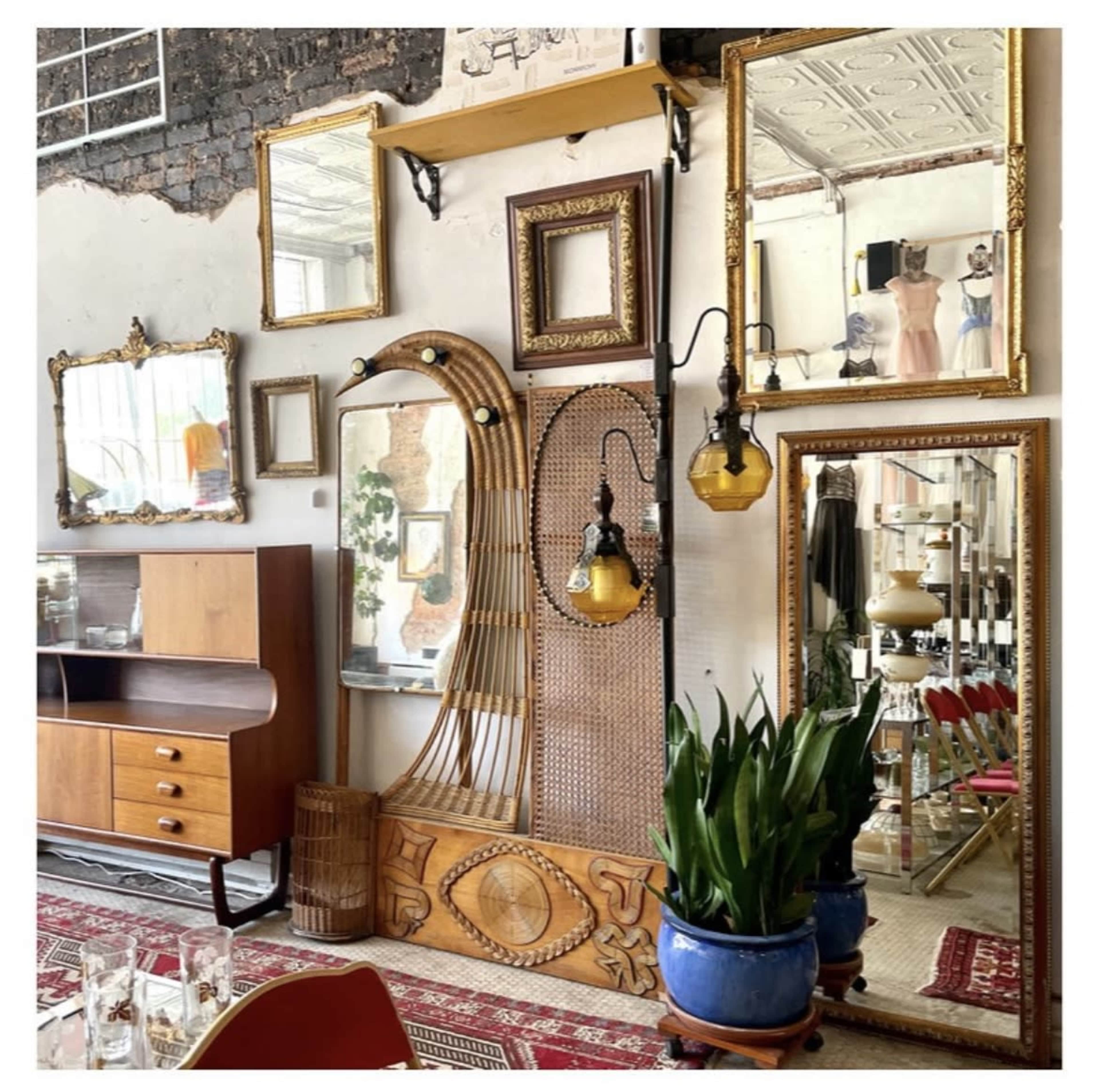 A vintage interior with a wooden furniture piece, multiple ornate mirrors, a decorative wall panel, plants in blue pots, and hanging light fixtures.