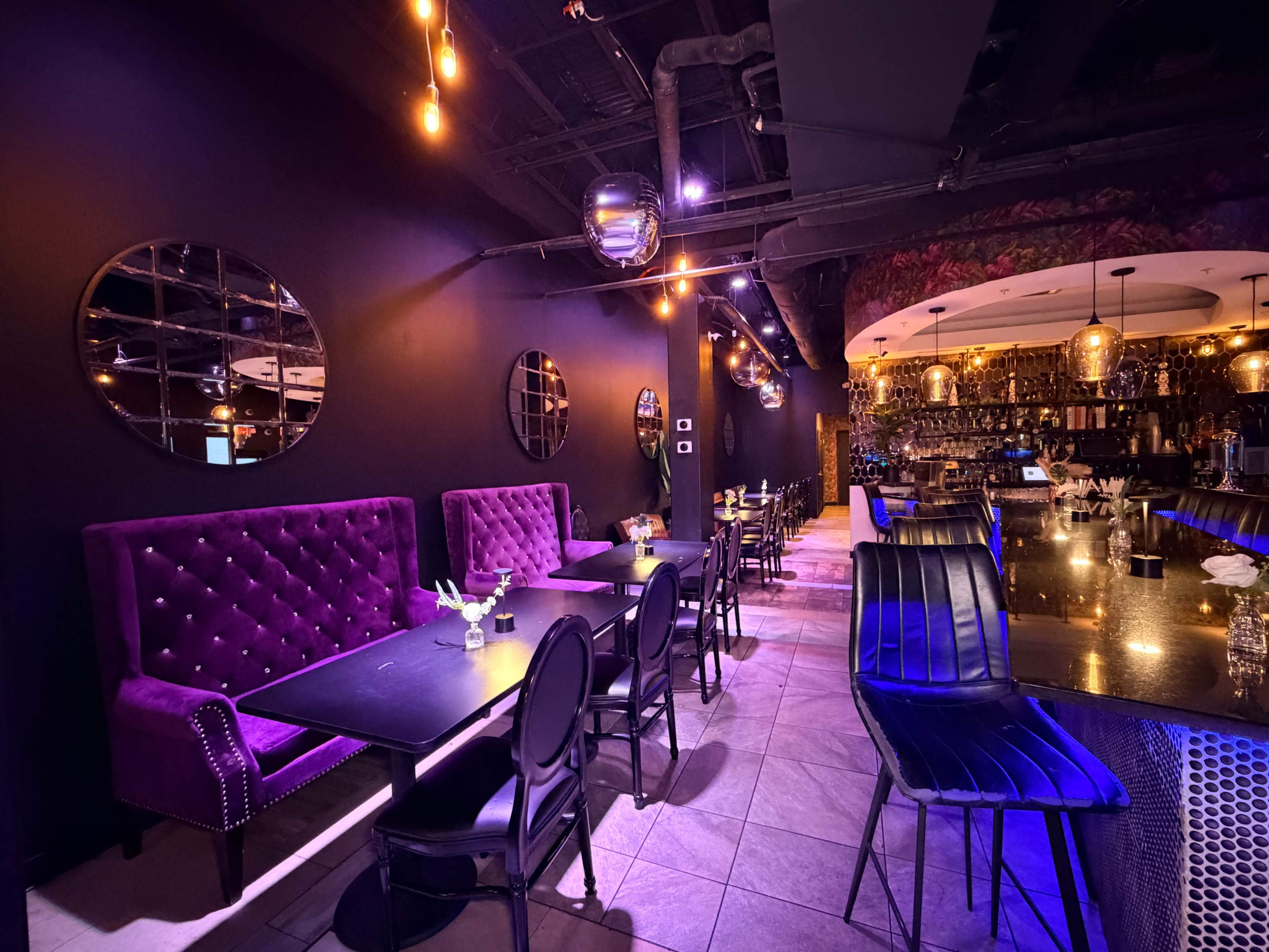 The image shows a stylish restaurant interior featuring purple upholstered booths, black tables, and modern lighting fixtures along a dark-colored wall.