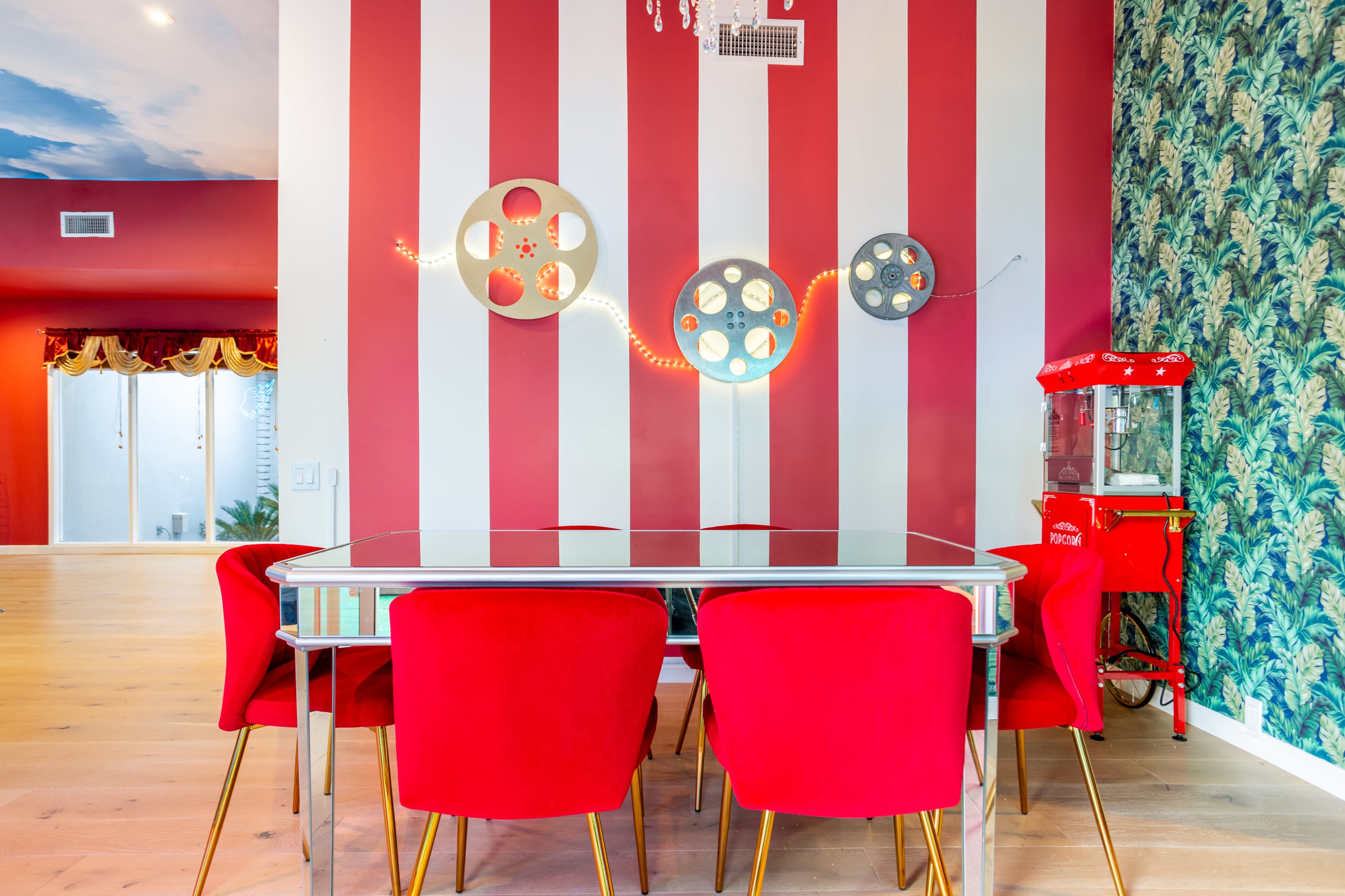 A dining area with a glass table surrounded by red chairs, set against striped walls and a textured wallpaper, with film reels and a vintage popcorn machine for decor.