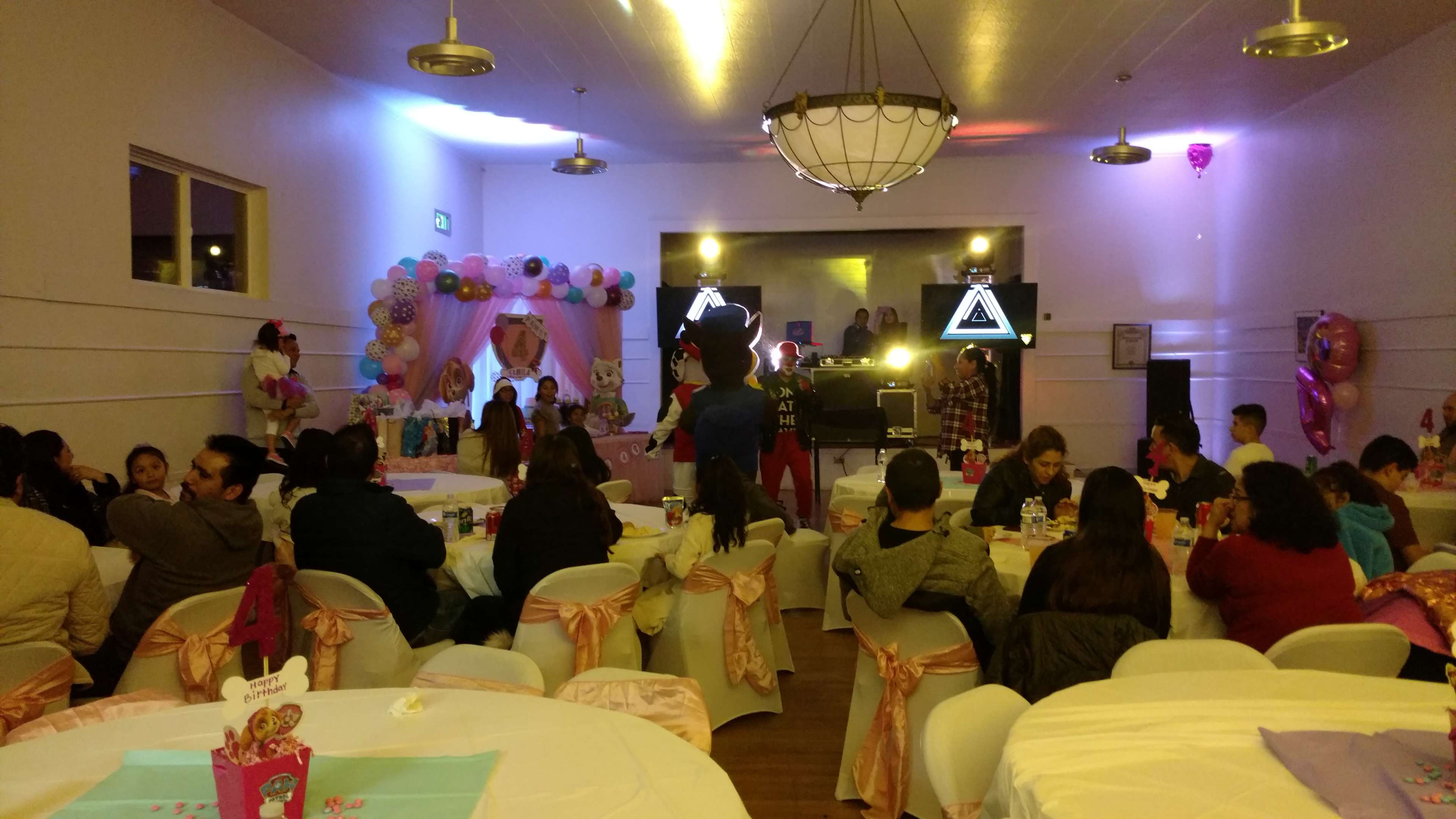 A party scene is set indoors, with tables adorned in pink and white decorations, featuring balloons and a stage with lighting effects in the background.