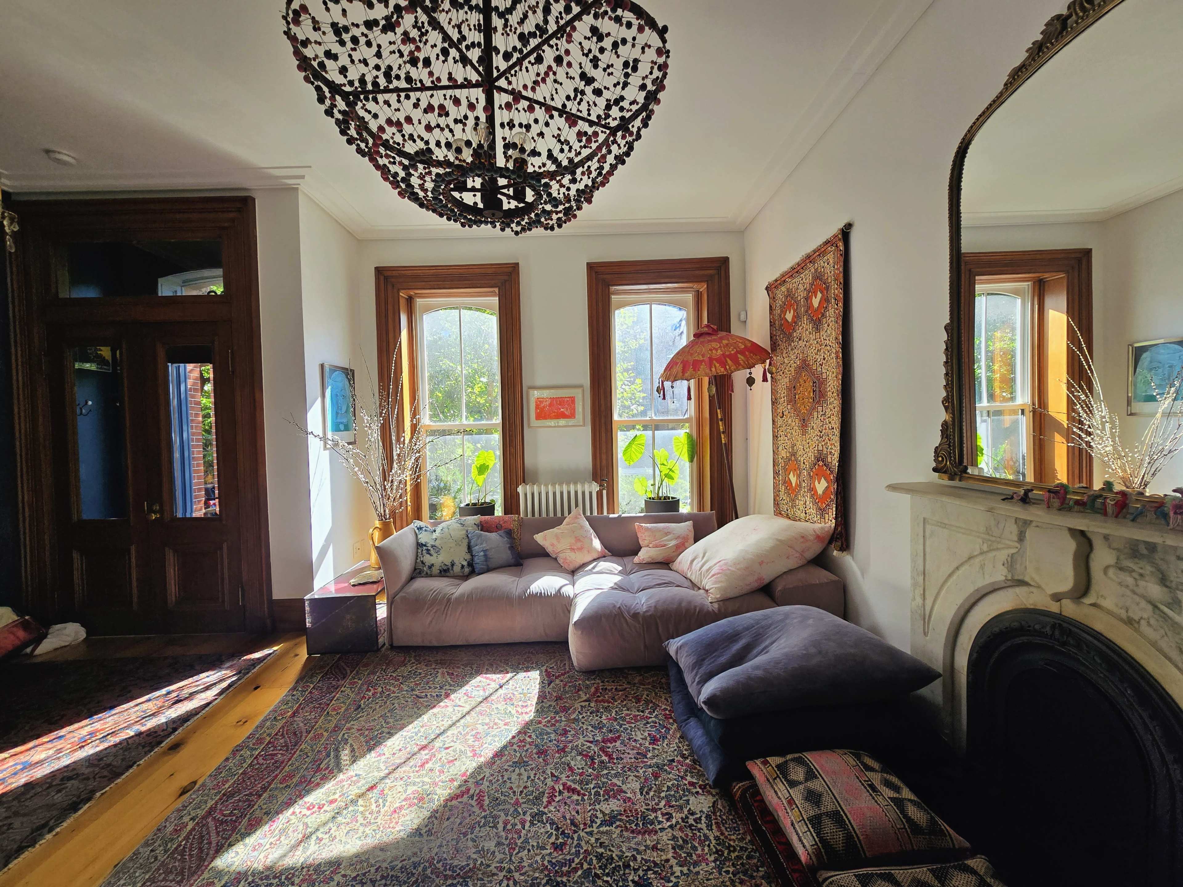 The image shows a cozy living room with large windows, a patterned area rug, a plush couch, and a decorative chandelier.