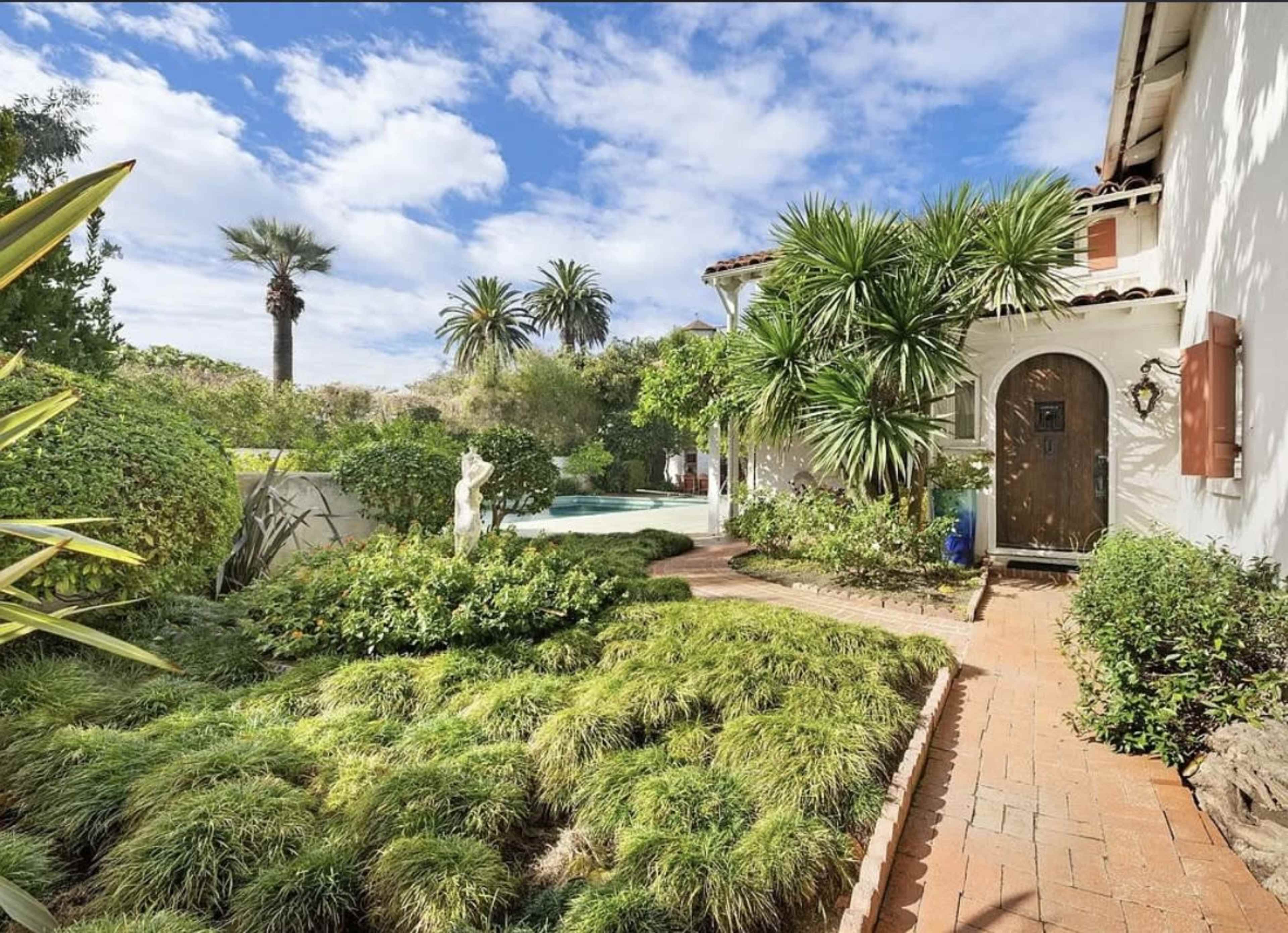 The image shows a landscaped garden with a stone pathway leading to a house, a swimming pool in the background, and palm trees and greenery surrounding the area.