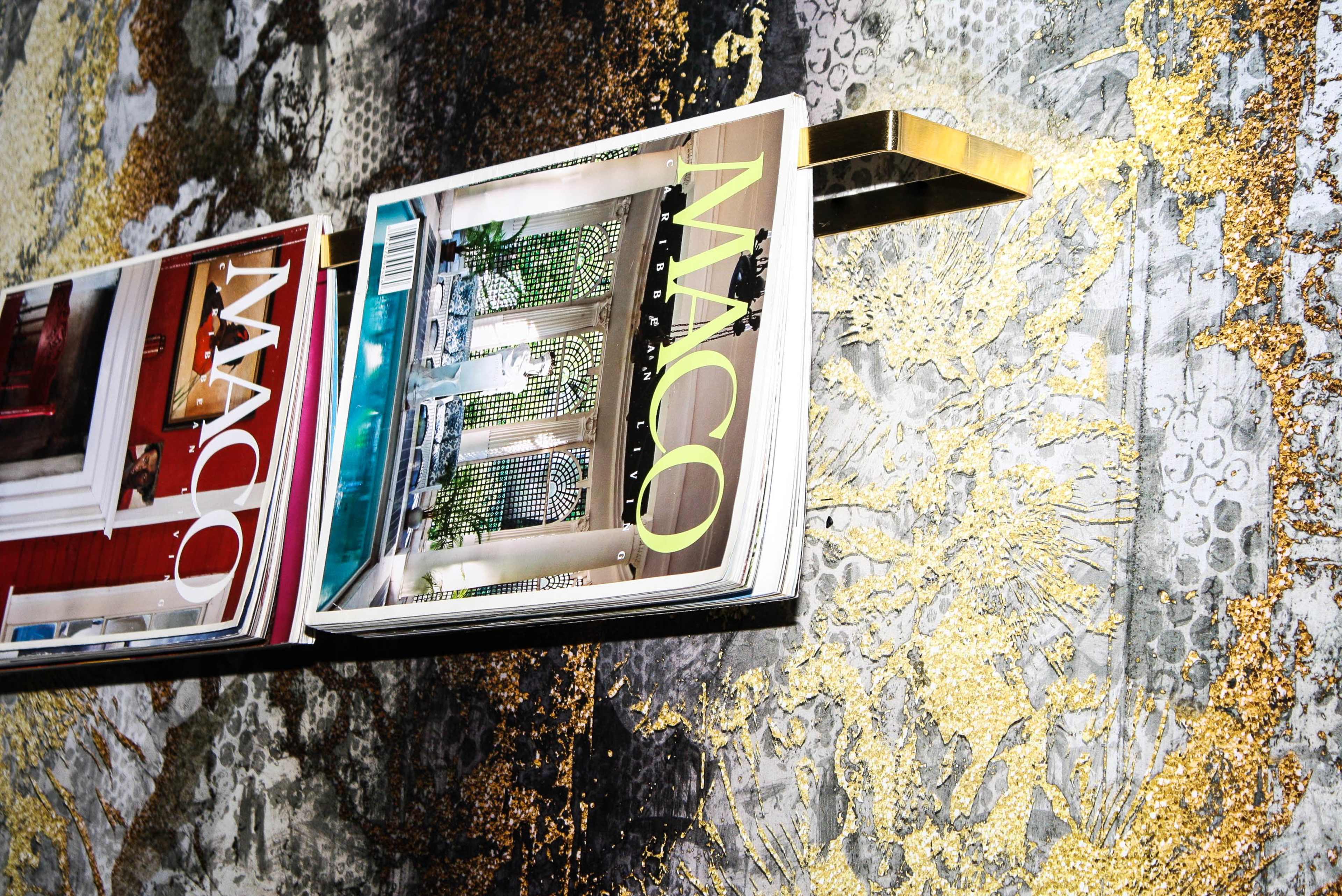 A metal rack holds magazines featuring interior design and architecture against a decorated wall with a gold pattern.