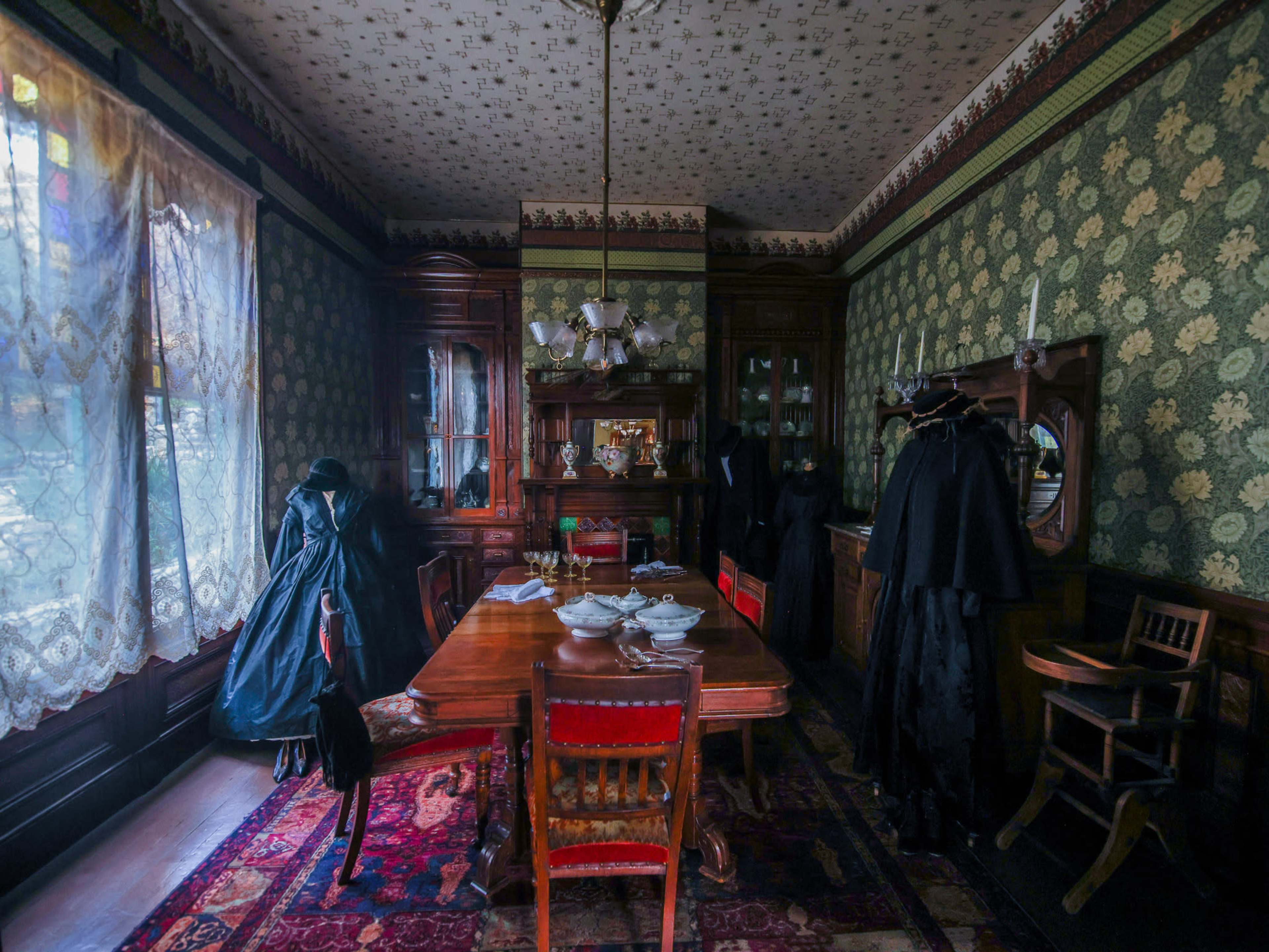 A darkly decorated dining room features a large wooden table set with dishes, surrounded by antique furniture and cloaked figures in long garments.