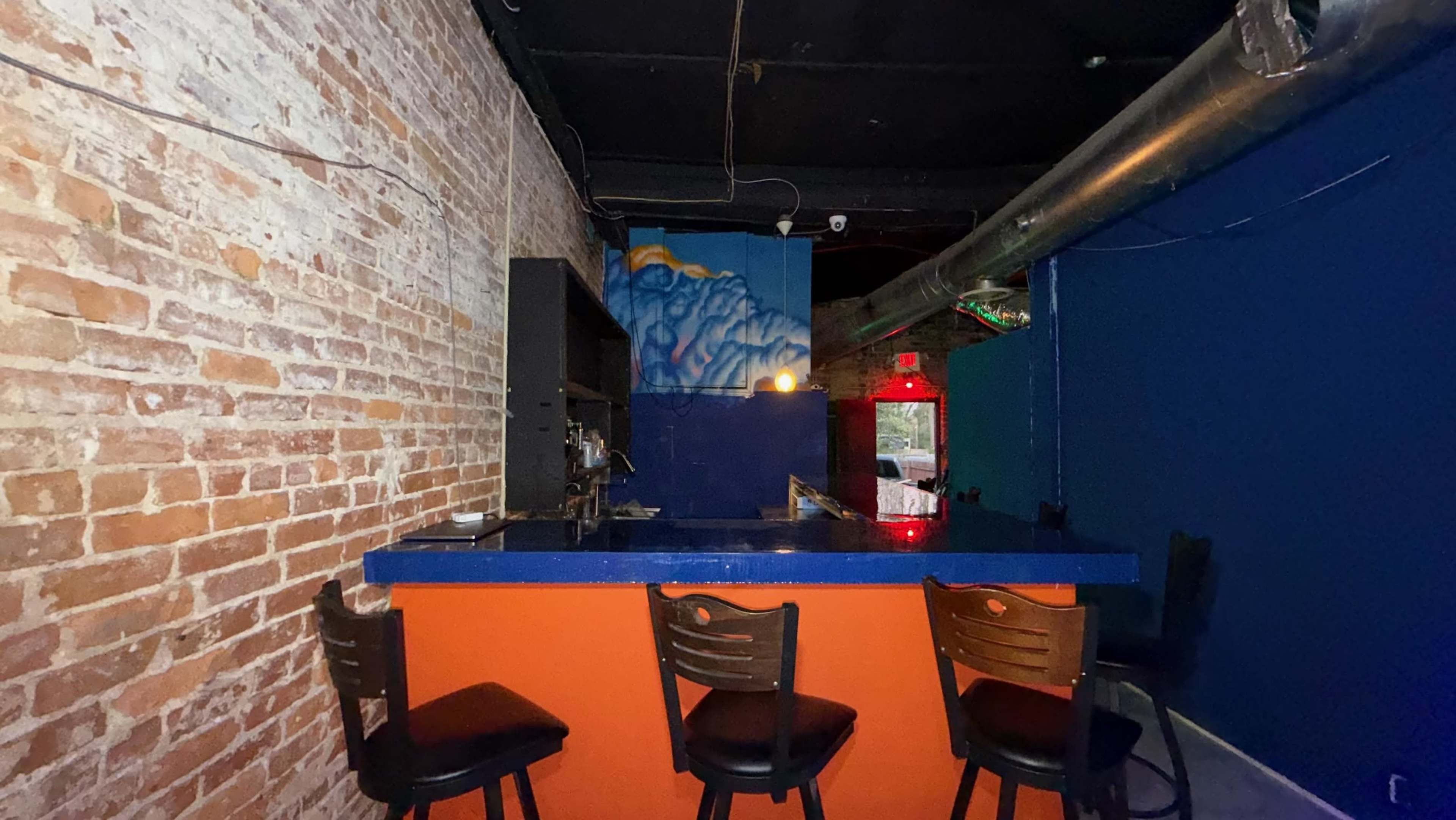 The image shows a small bar area with a blue and orange counter, wooden stools, and exposed brick walls.