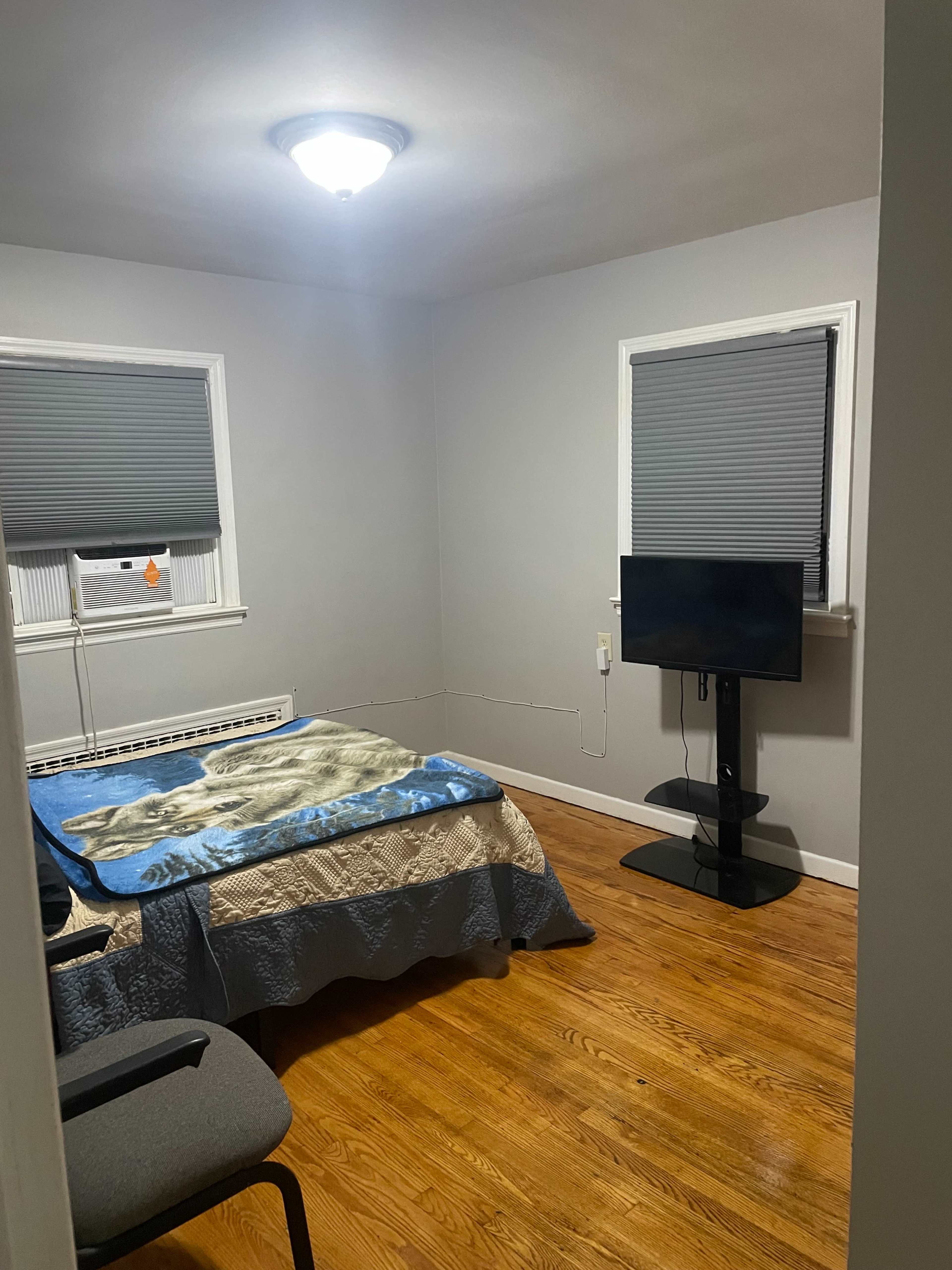 The image shows a simple bedroom with a bed covered in a blue blanket, a television mounted on a stand, and wooden flooring.
