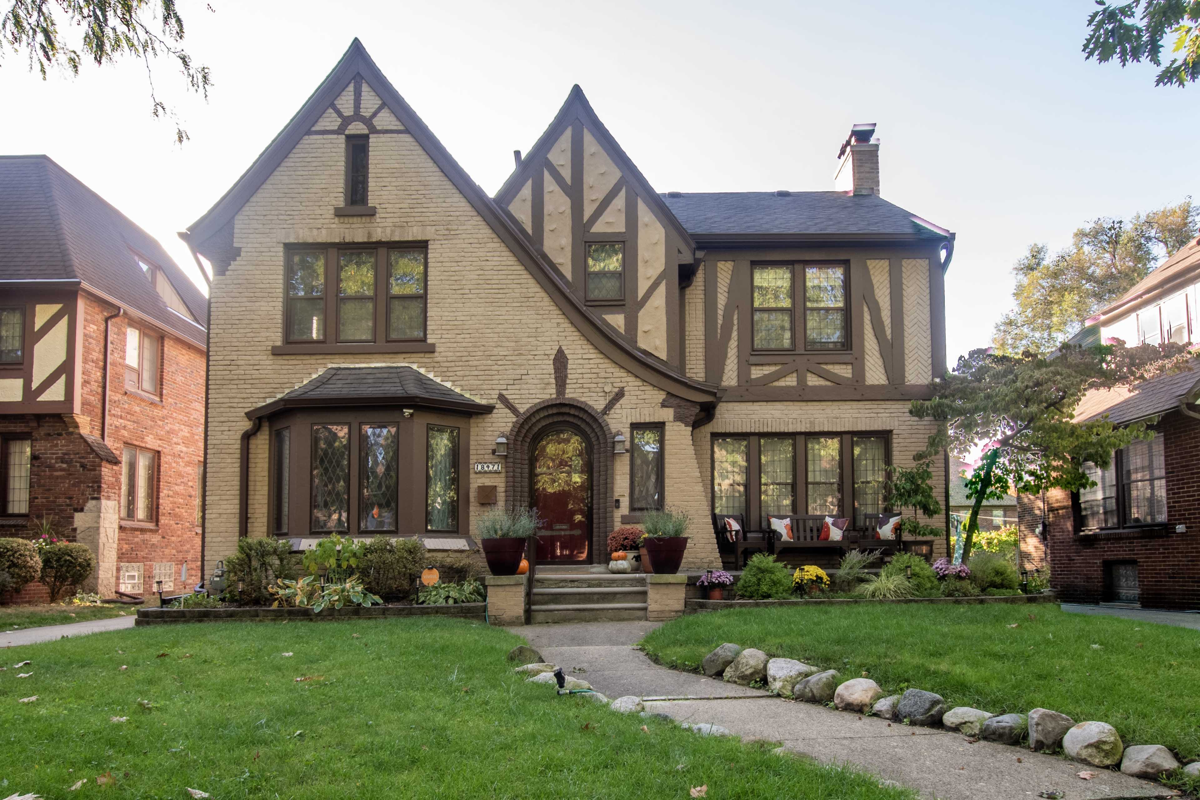 The image shows a two-story Tudor-style house with a brick and stucco exterior, a central front door, and a landscaped yard featuring greenery and flowers.