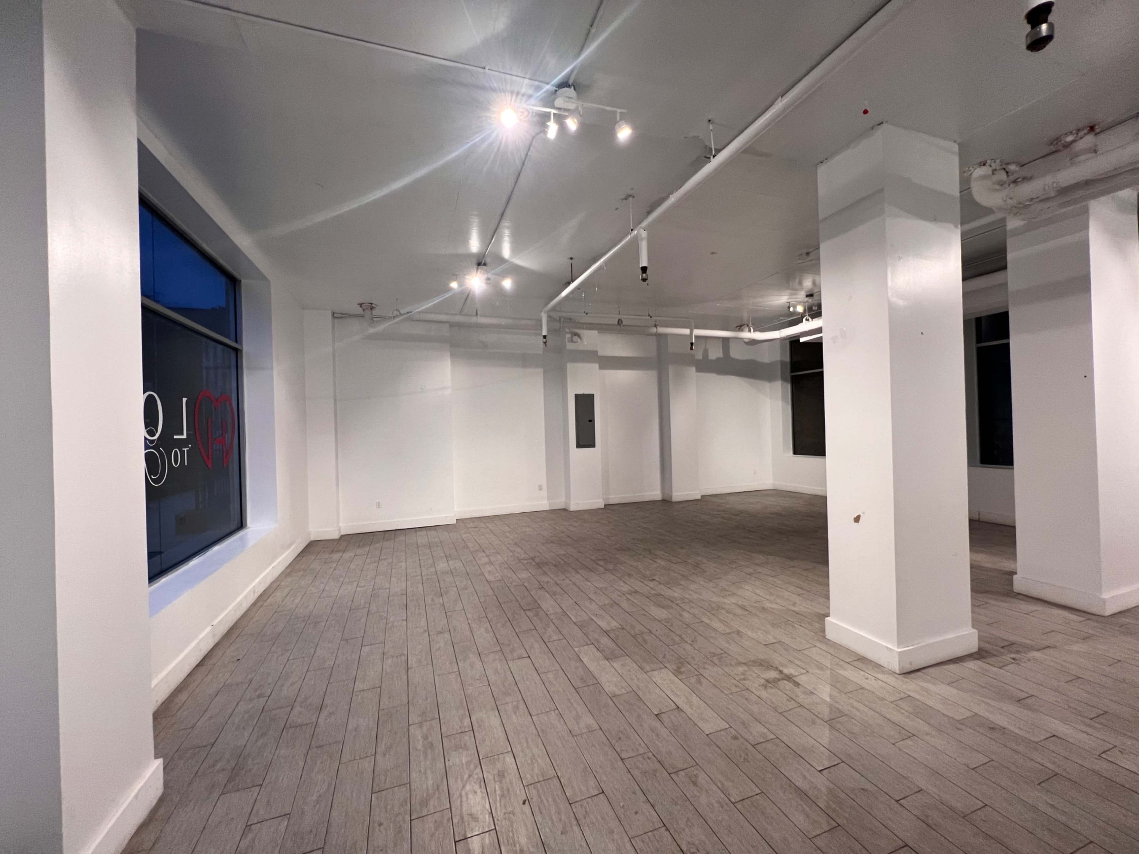 An empty, well-lit room with wooden flooring and white walls, featuring several overhead lights and large windows.