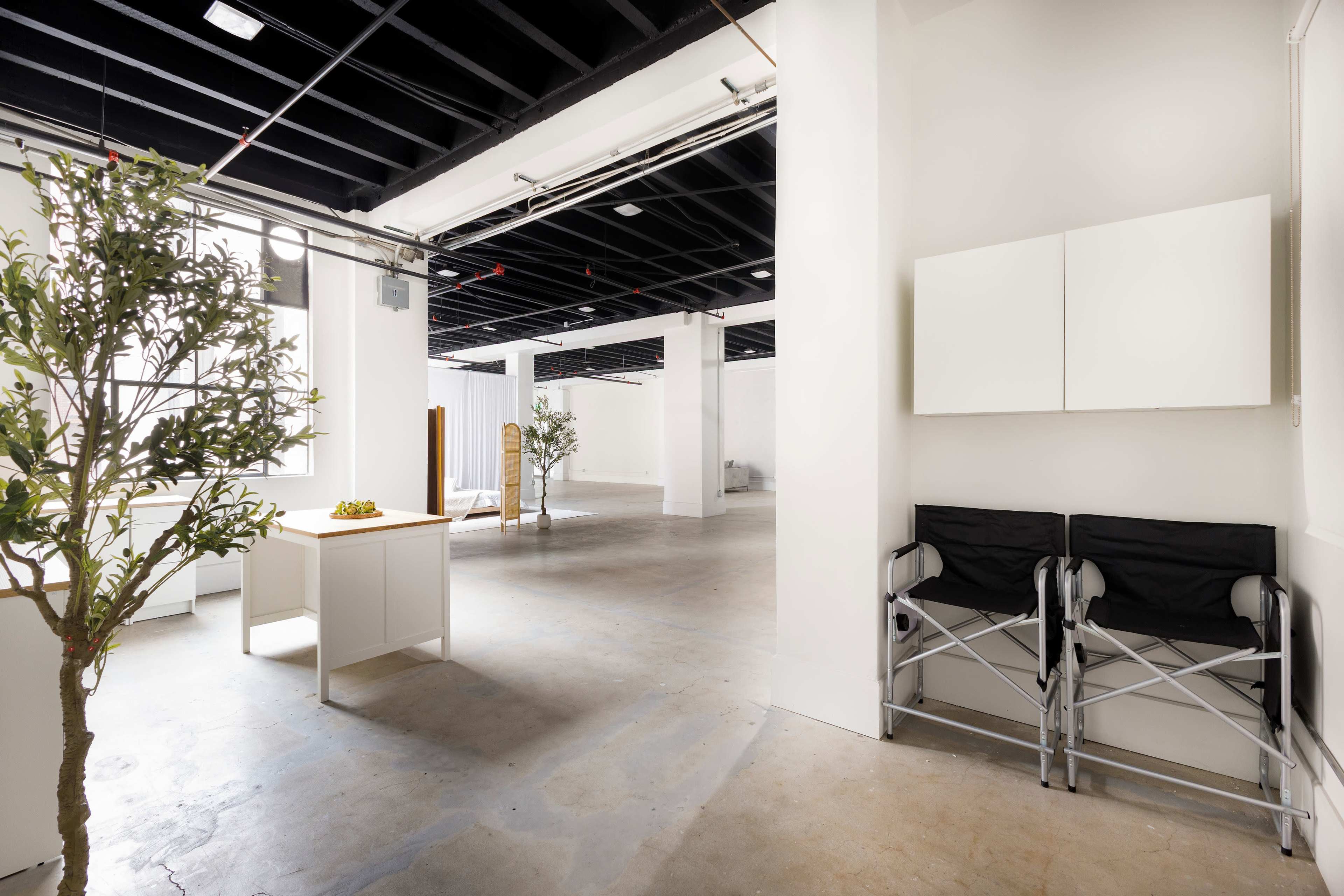 An empty room featuring a minimalist design with two folding chairs, a small table holding fruit, and a couple of potted trees.