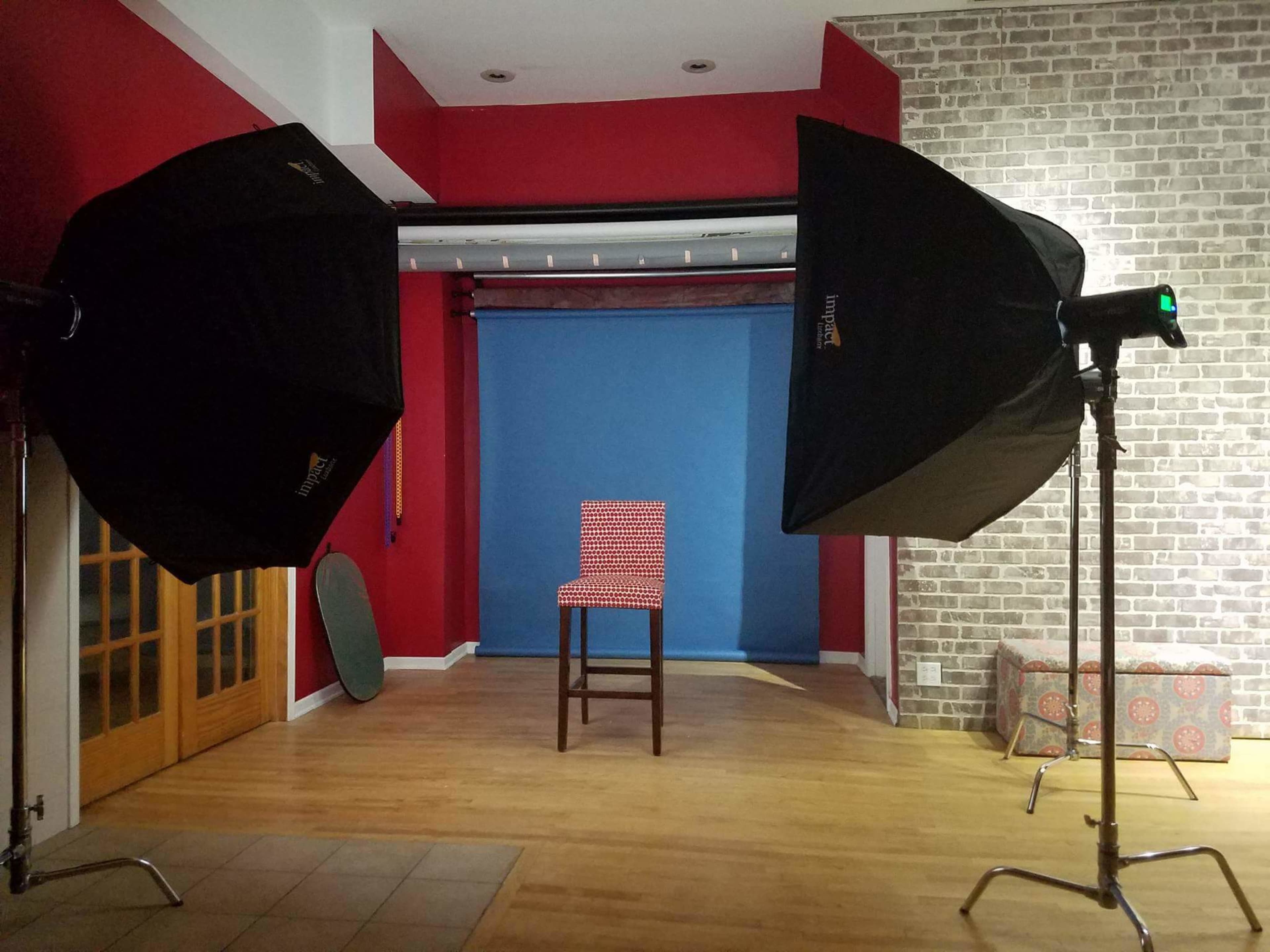 A photography studio is set up with two large softbox lights illuminating a red and white checkered stool against a blue backdrop.