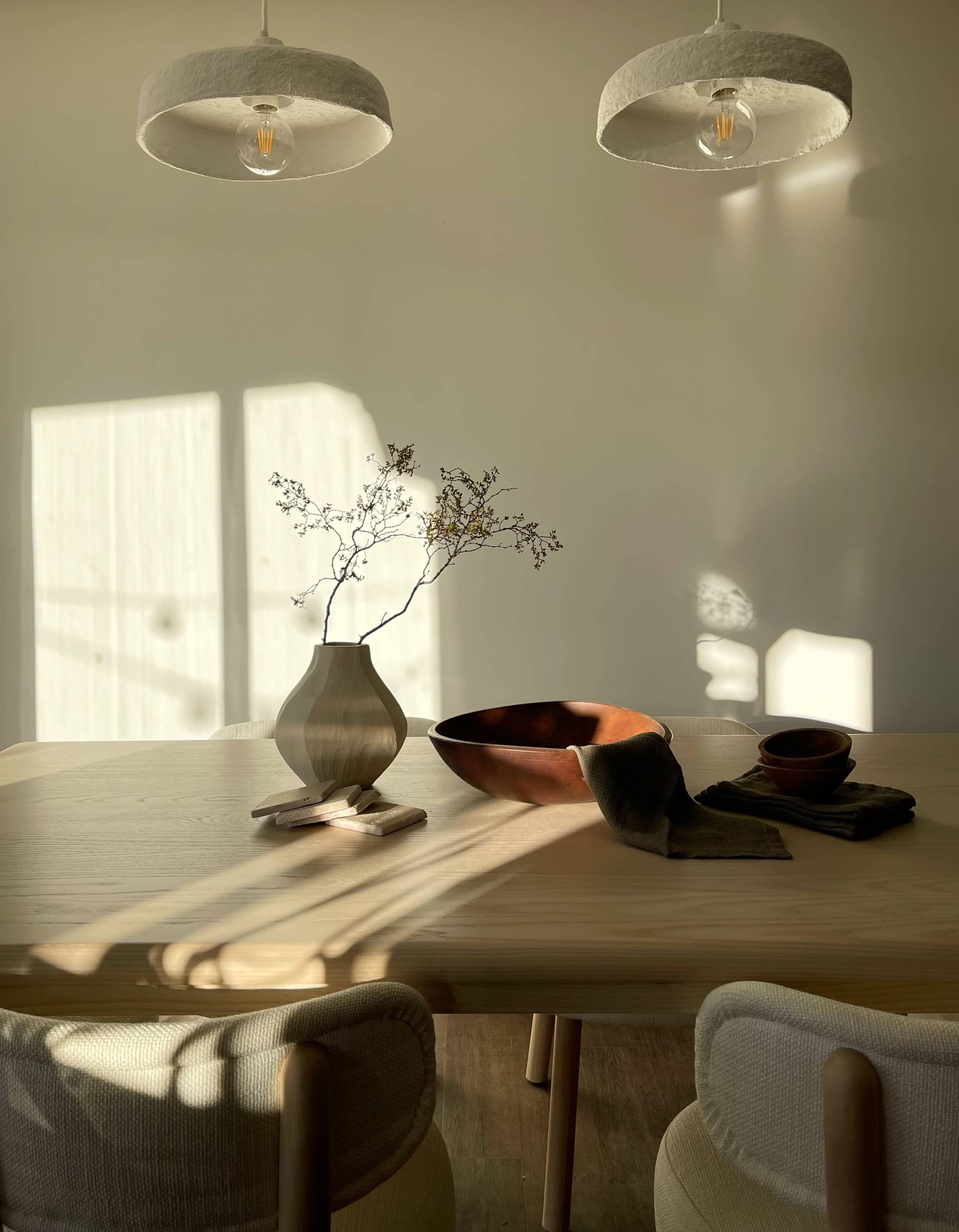 A minimalist dining table set with a wooden bowl, a ceramic vase containing branches, and textile items, illuminated by sunlight casting shadows from two pendant lamps.