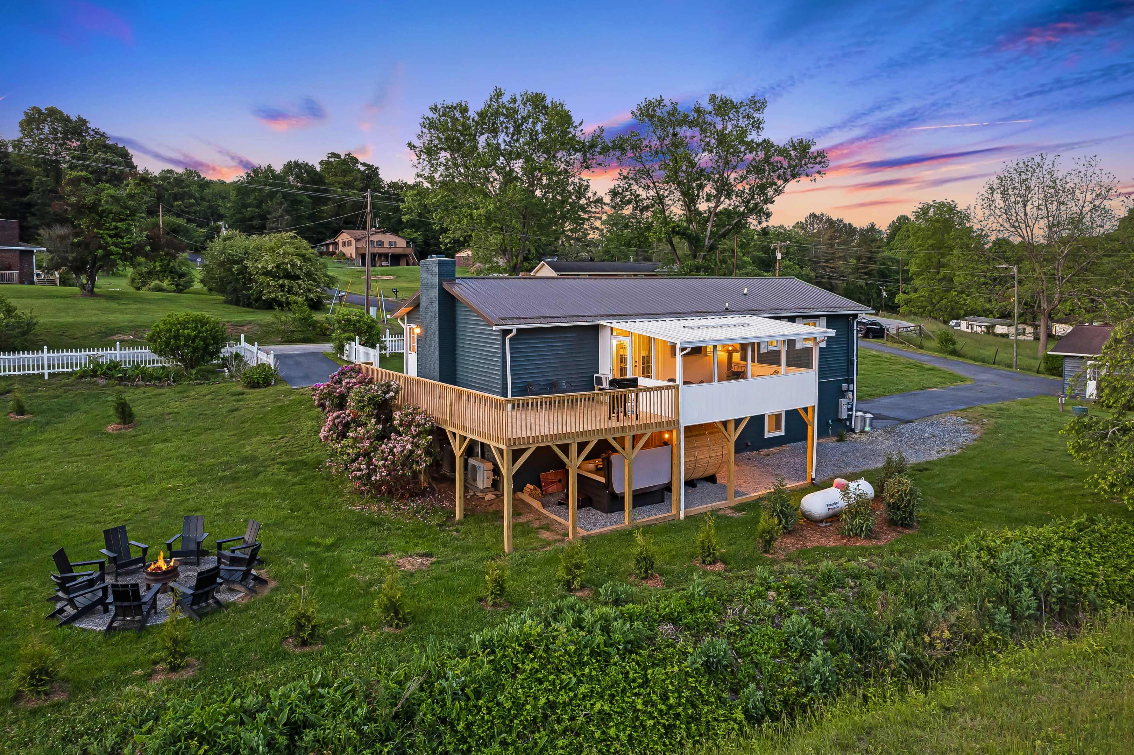 The image shows a two-story blue house with a wooden deck overlooking a green lawn and a fire pit surrounded by seating in a rural setting at sunset.