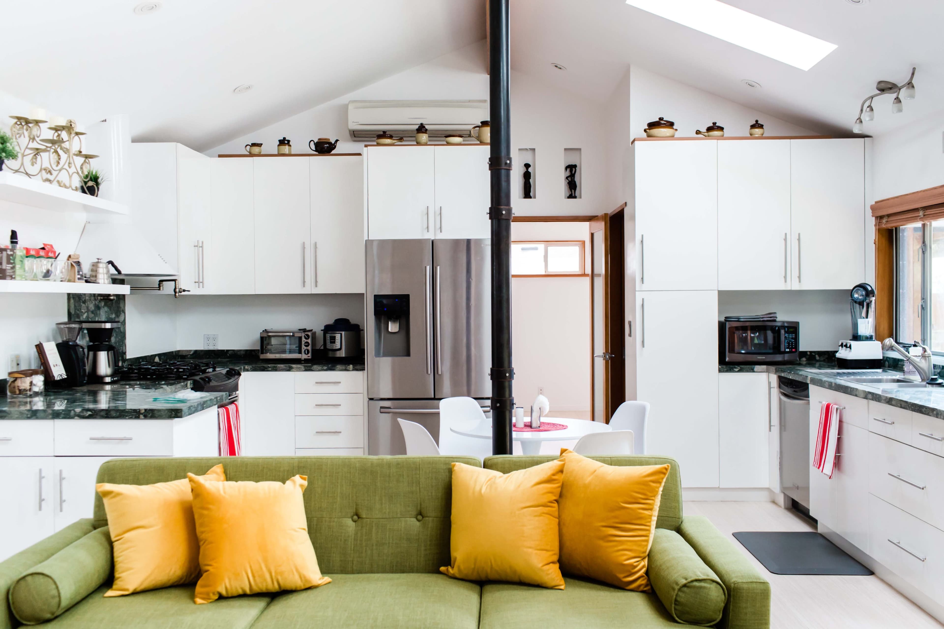 A modern kitchen features white cabinetry and a green sofa with yellow pillows, with a dining area visible in the background.