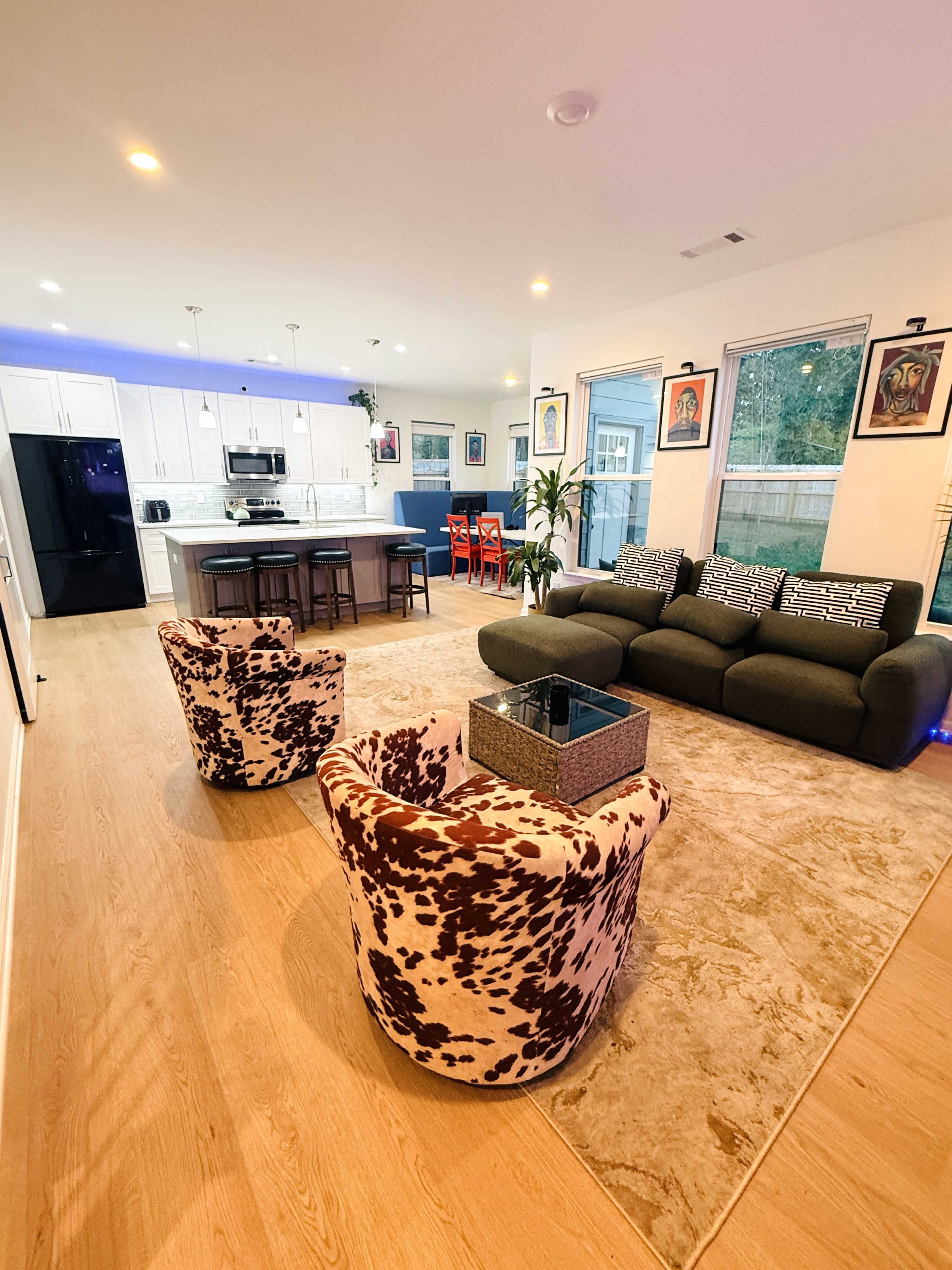 The image shows a modern living room and kitchen area featuring a large rug, two patterned chairs, a sectional sofa, and a dining table with colorful chairs.