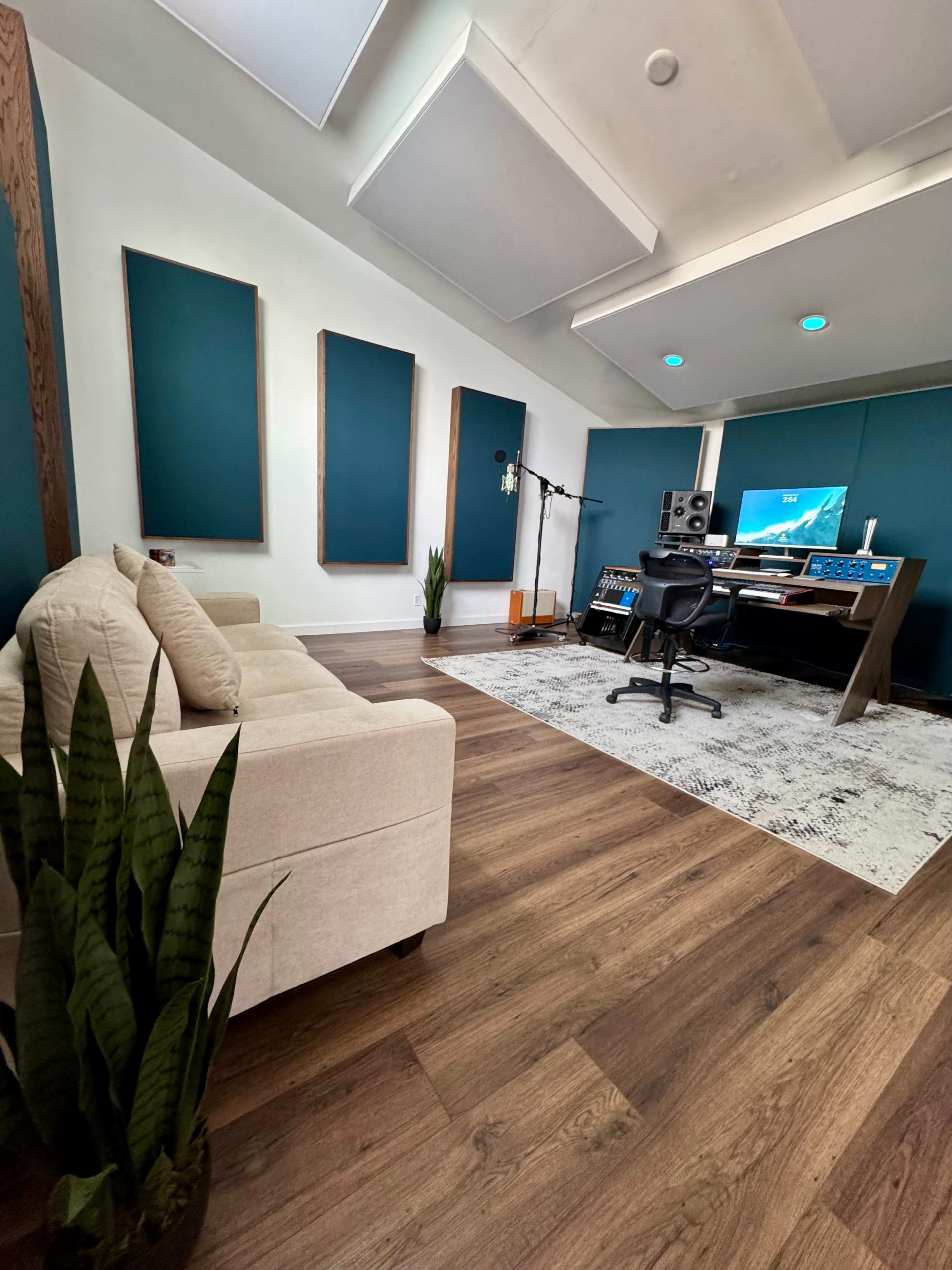 A modern recording studio features a desk with audio equipment, a black chair, a couch, and soundproof panels on the walls.