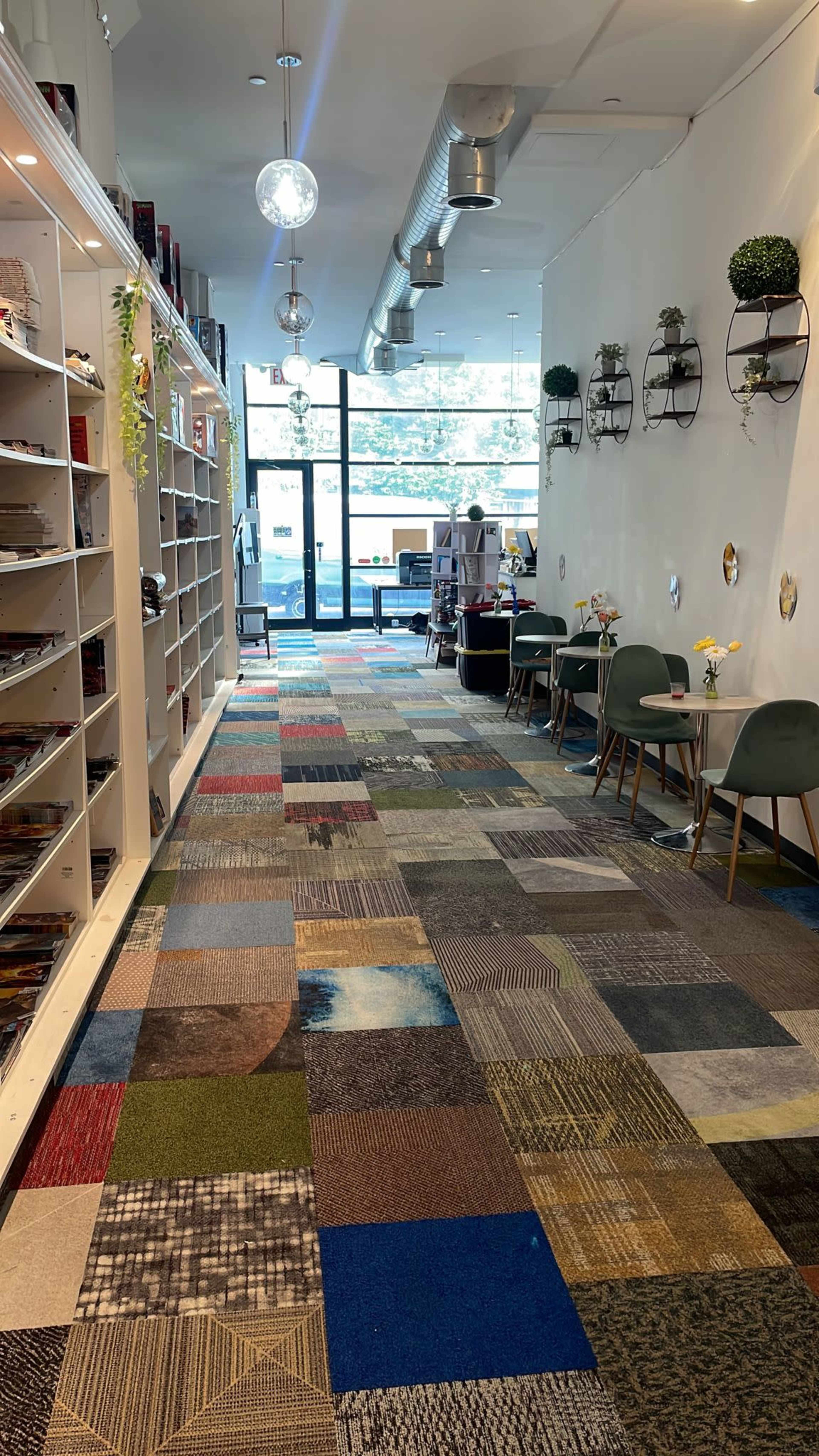 A bright corridor with shelves of books on one side and tables with chairs on the other, featuring a multi-colored patchwork carpet.
