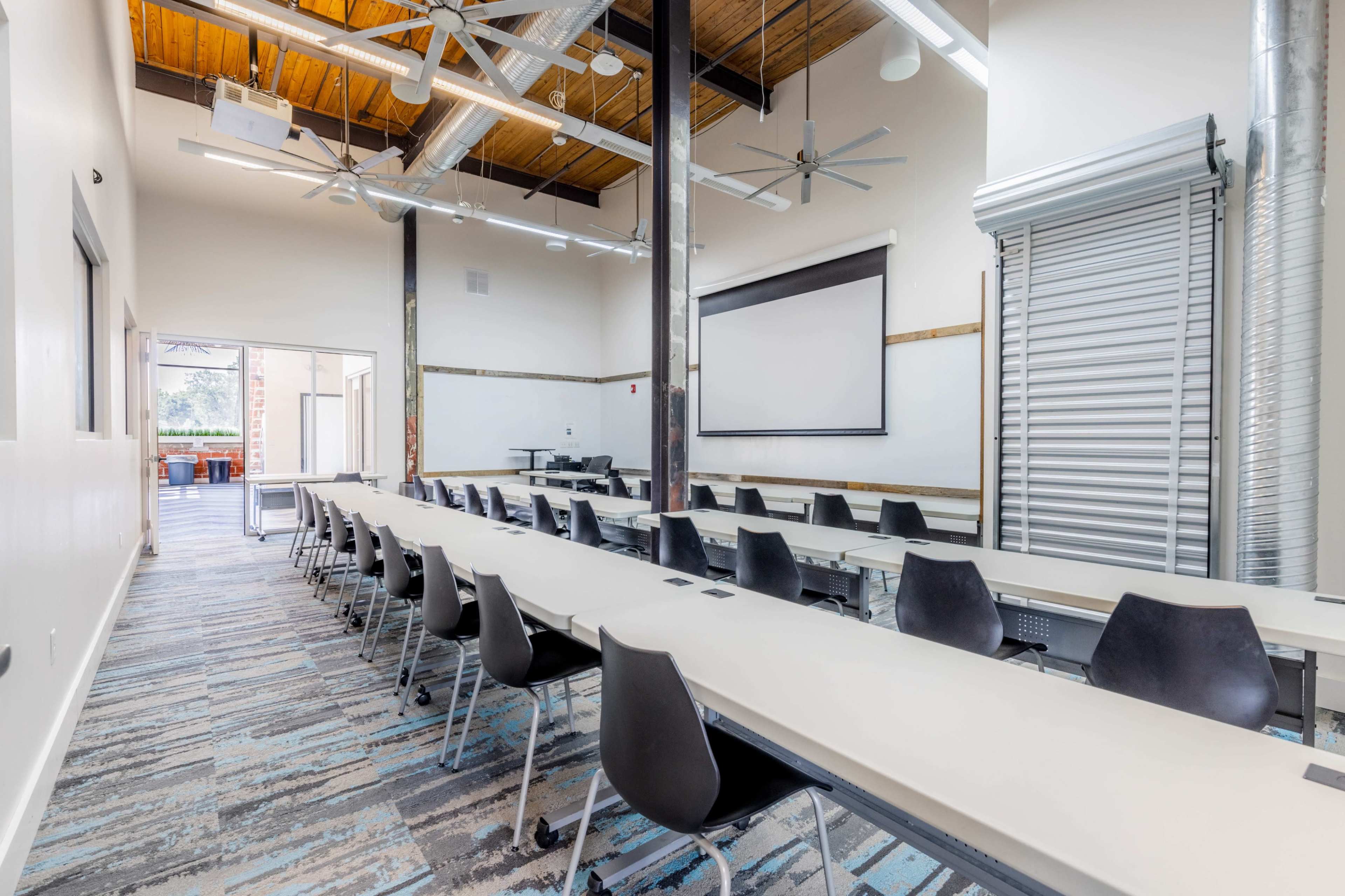 The image shows a modern classroom with long tables and black chairs, featuring a projector screen and large windows that open to an outdoor area.