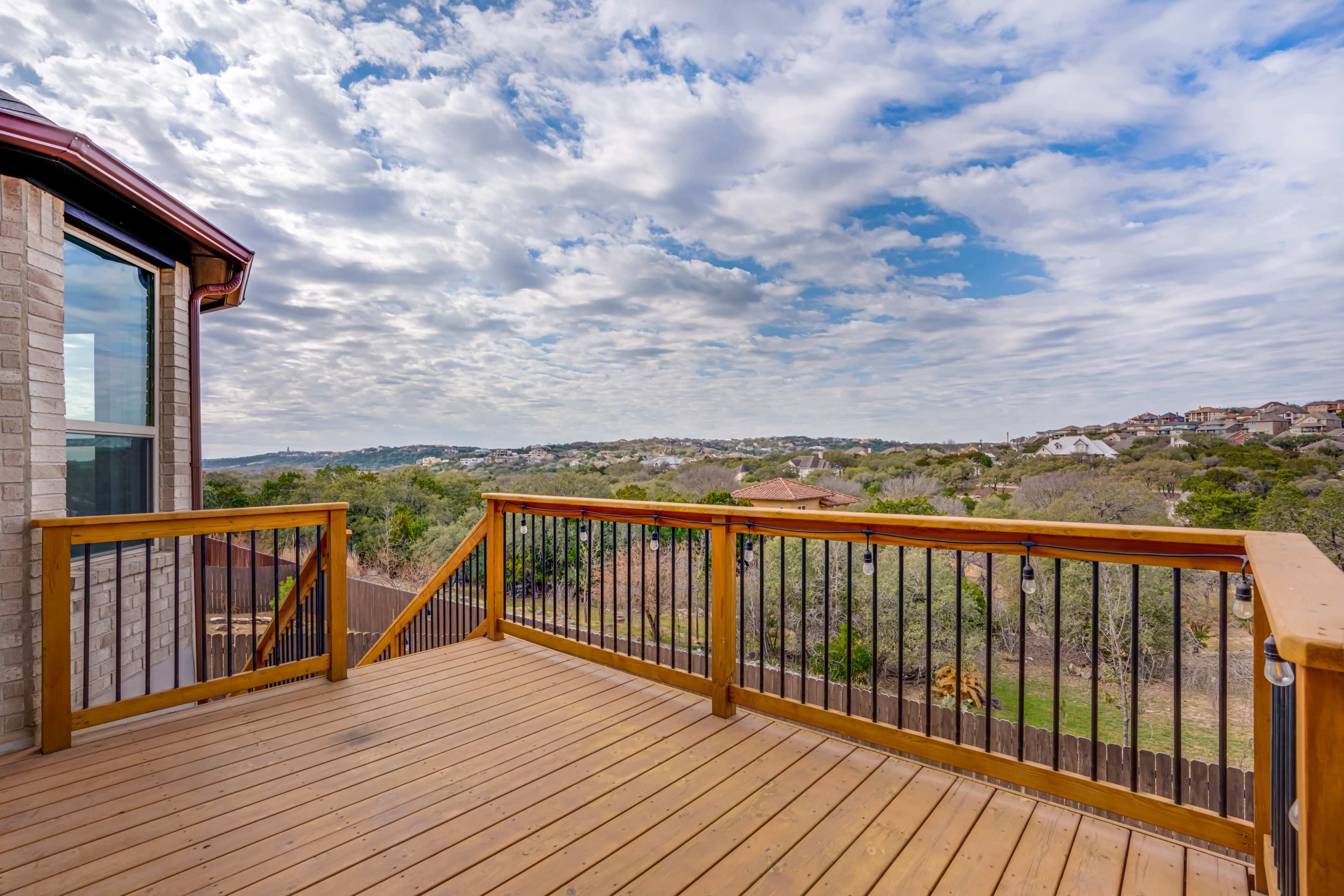 A wooden deck with a railing overlooks a rural landscape dotted with trees and distant houses under a cloudy sky.