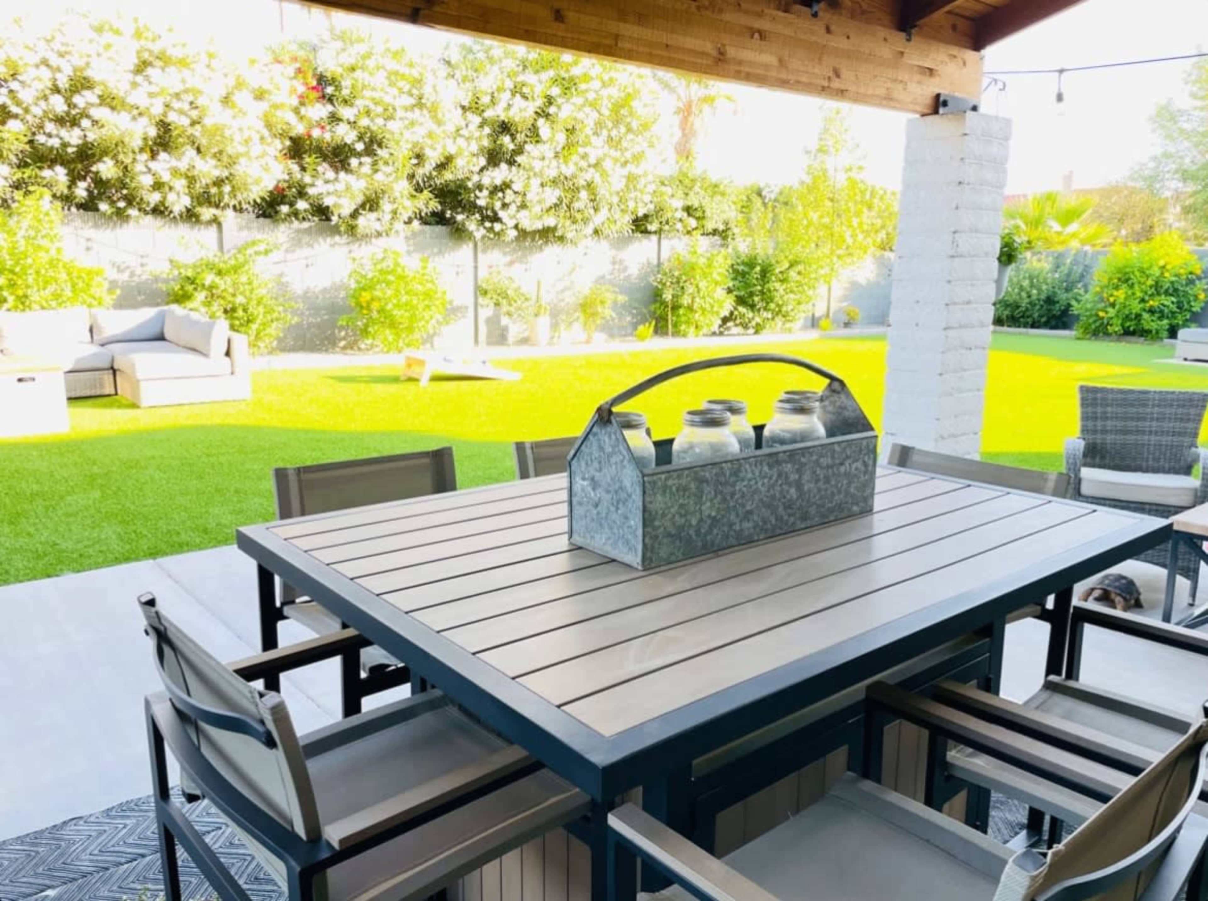 A metal table with a wooden surface and six chairs is set up on a patio surrounded by greenery and a well-maintained lawn.
