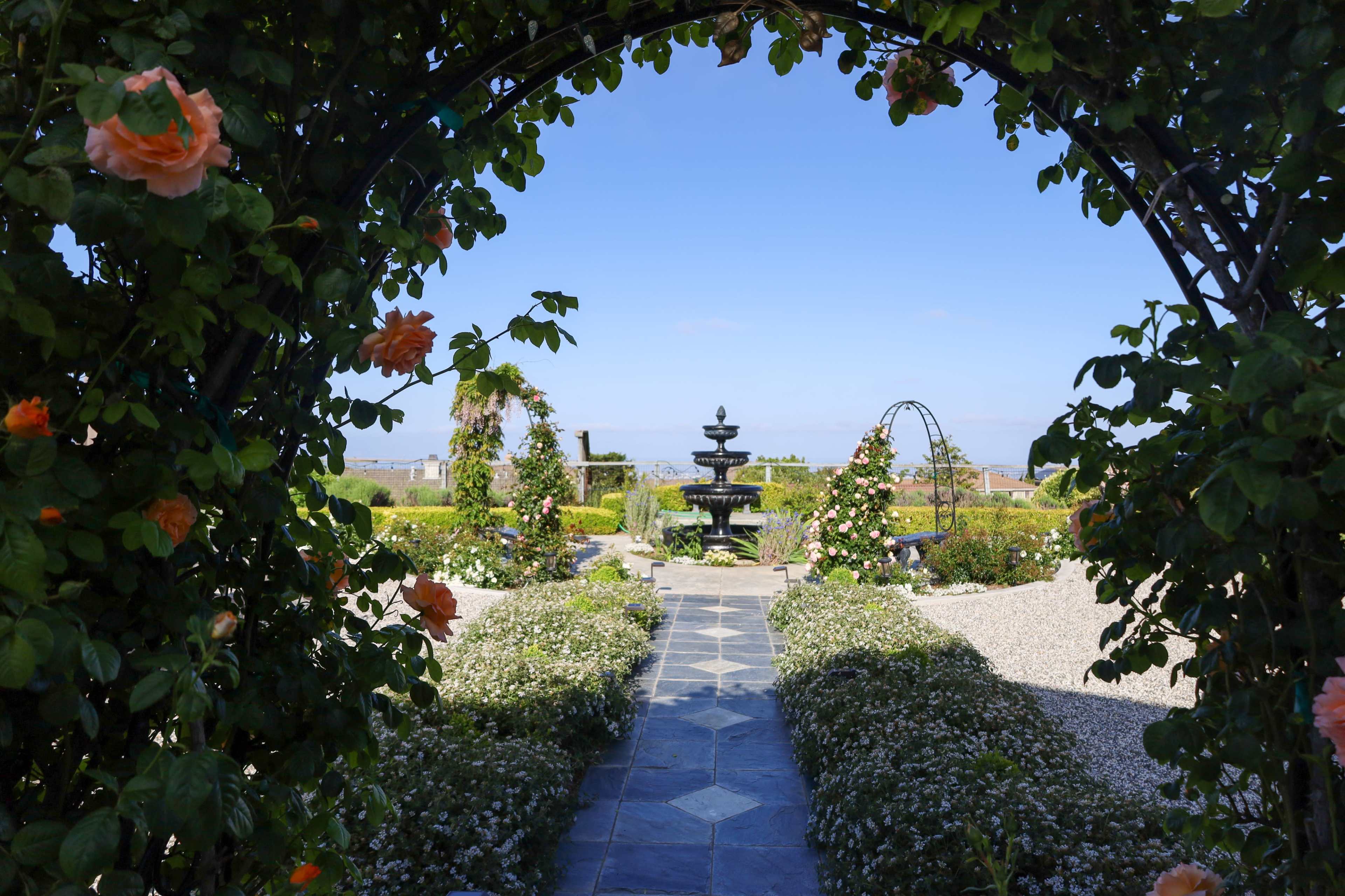 A stone pathway leads through an archway adorned with climbing roses to a central fountain surrounded by gardens.